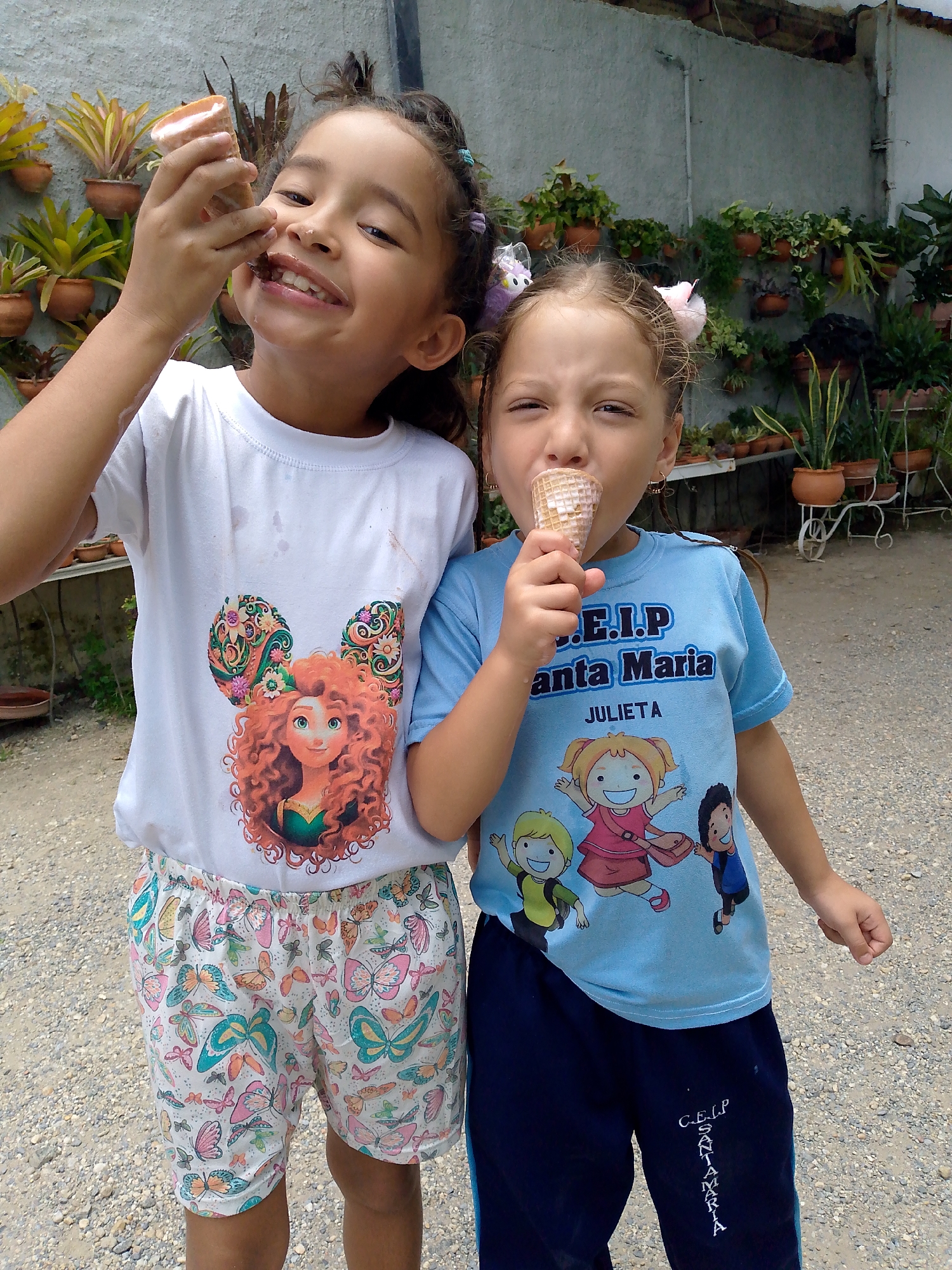Two young girls stand outdoors, each holding and eating an ice cream cone. The girl on the left is fair-skinned and has curly brown hair. She wears a white t-shirt with a Disney's "Brave" character design on it, paired with patterned shorts. The girl on the right is also fair-skinned with brown hair, adorned with hair accessories. She wears a light blue t-shirt with the inscription "C.E.I.P Santa Maria JULIETA" and a cartoon graphic. She is wearing blue trousers with the name of the school C.E.I.P SANTA MARIA printed. The background includes a wall covered with potted plants, suggesting a garden or courtyard setting. The ground is covered with gravel. The image was taken in Tinaquillo, Venezuela.