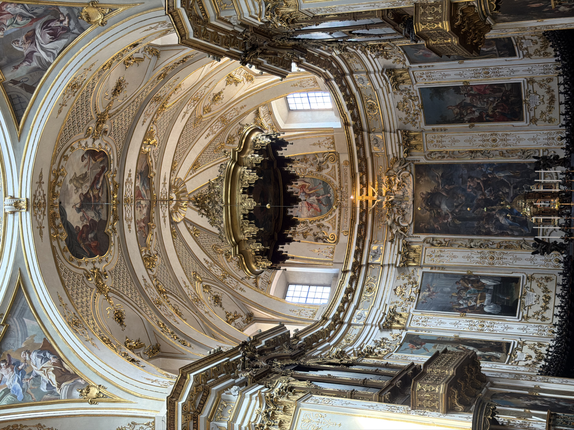 This is a photograph of the interior of a church, likely the Duomo di Mozzo, located in Mozzo, Italy. The image showcases the opulent baroque architecture of the ceiling and apse. The ceiling is intricately adorned with frescoes and gilded decorative elements, featuring detailed stucco work and ornate gold trim along the arched vaults. A large, elaborate chandelier hangs from the central dome. The walls of the apse are decorated with large oil paintings depicting biblical scenes, framed by gilded architectural details. Two large pipe organs are visible on either side of the apse, their ornate casings made of dark wood and gilded elements. Numerous windows with arched tops are present in the upper sections of the apse, allowing natural light to filter into the space. The overall impression is one of grandeur and religious devotion, typical of historical Italian cathedrals.