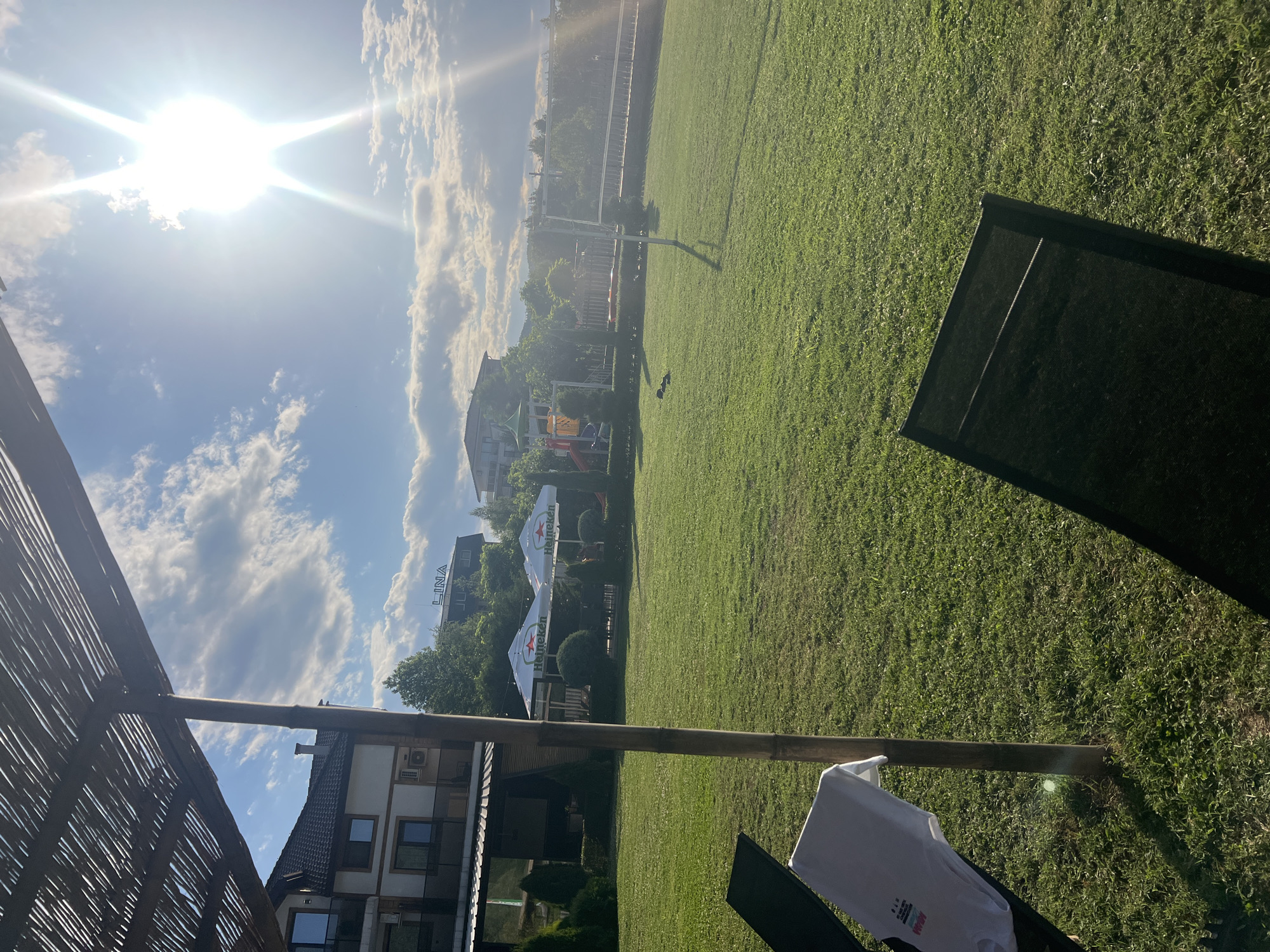 A sun-drenched, outdoor recreational area is depicted, likely in Kirkovo, Bulgaria. In the foreground, a portion of a thatched roof structure and a wooden pole are visible, casting shadows on a neatly mowed green lawn. Two lounge chairs are placed on the grass, one with a white t-shirt draped over it. In the middle ground, two large Heineken-branded umbrellas provide shade over tables and chairs. Beyond this, a playground with colorful equipment is situated. In the background, several multi-story buildings are visible, suggesting a hotel or residential complex. The sky is a vibrant blue with scattered white clouds, and the sun is intensely bright, creating lens flares and a hazy atmosphere. The overall scene conveys a sense of leisure and relaxation.