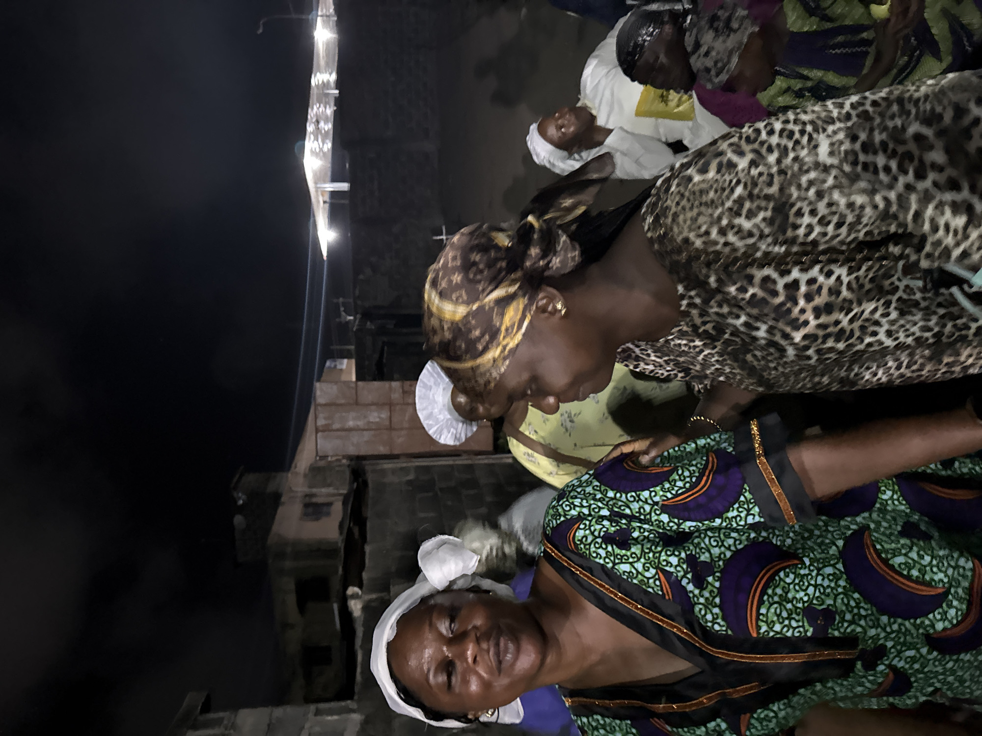 The image shows a group of African women outdoors in what appears to be a night scene. The woman in the foreground is wearing a green, blue, and purple patterned dress with a white head covering. The woman behind her is wearing a brown leopard-print top and a head wrap. A third woman is wearing a white head covering and a yellow shirt. The background shows a building with lights. The image's location is presumed to be Badagry, Nigeria, based on the prompt's instructions.