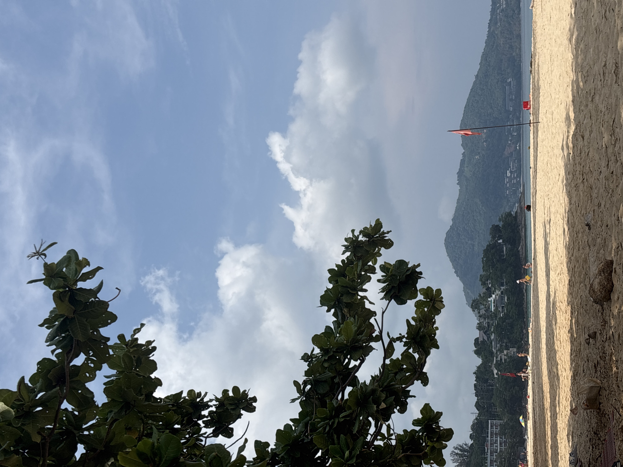 The image depicts a beach scene in Choeng Thale, Thailand. A tree with dark green foliage occupies the lower left corner of the frame. In the background, a beach stretches towards a headland covered in dense vegetation and buildings. Two red flags are visible, indicating potential hazards in the water. Several people are present on the beach. The sky is light blue with scattered white clouds. Sand makes up the right side of the image. The photo appears to be taken in daylight.