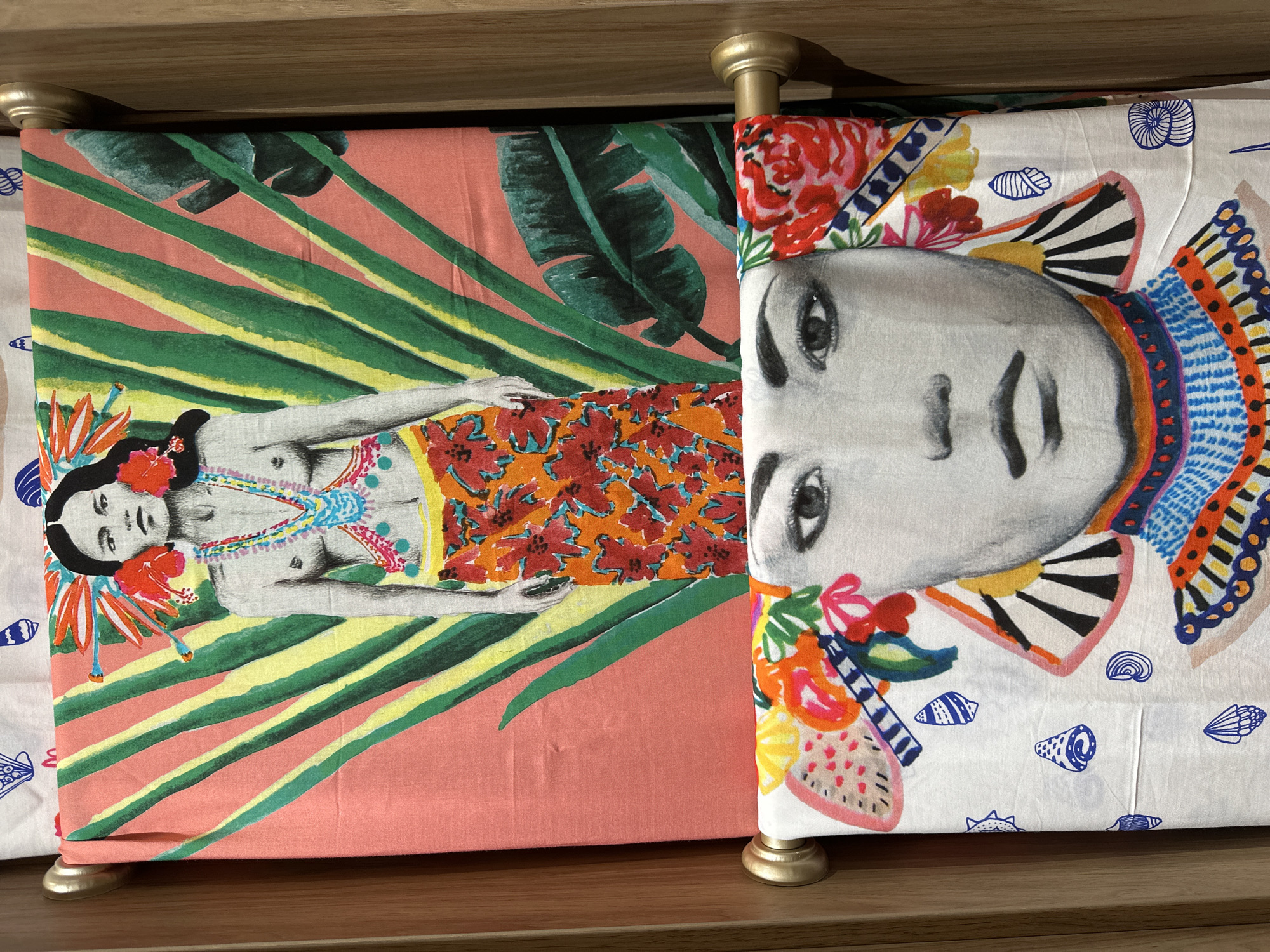 The image shows two fabric panels hanging vertically. The top panel features a stylized illustration of a woman with dark hair, adorned with bright red flowers and elaborate jewelry. She is depicted with a naked torso and is wearing a vibrant orange skirt with a floral pattern. Behind her, there are large, stylized green leaves and pink and yellow stripes.
The bottom panel displays a close-up black and white illustration of a woman's face. She has dark eyes, dark eyebrows, and dark lips. She is decorated with colorful, abstract patterns and flowers around her face. The fabric beneath her face is white and patterned with small blue shell designs. Both panels are draped over golden rods, which are attached to wooden supports.
The location is Kuta, Indonesia.