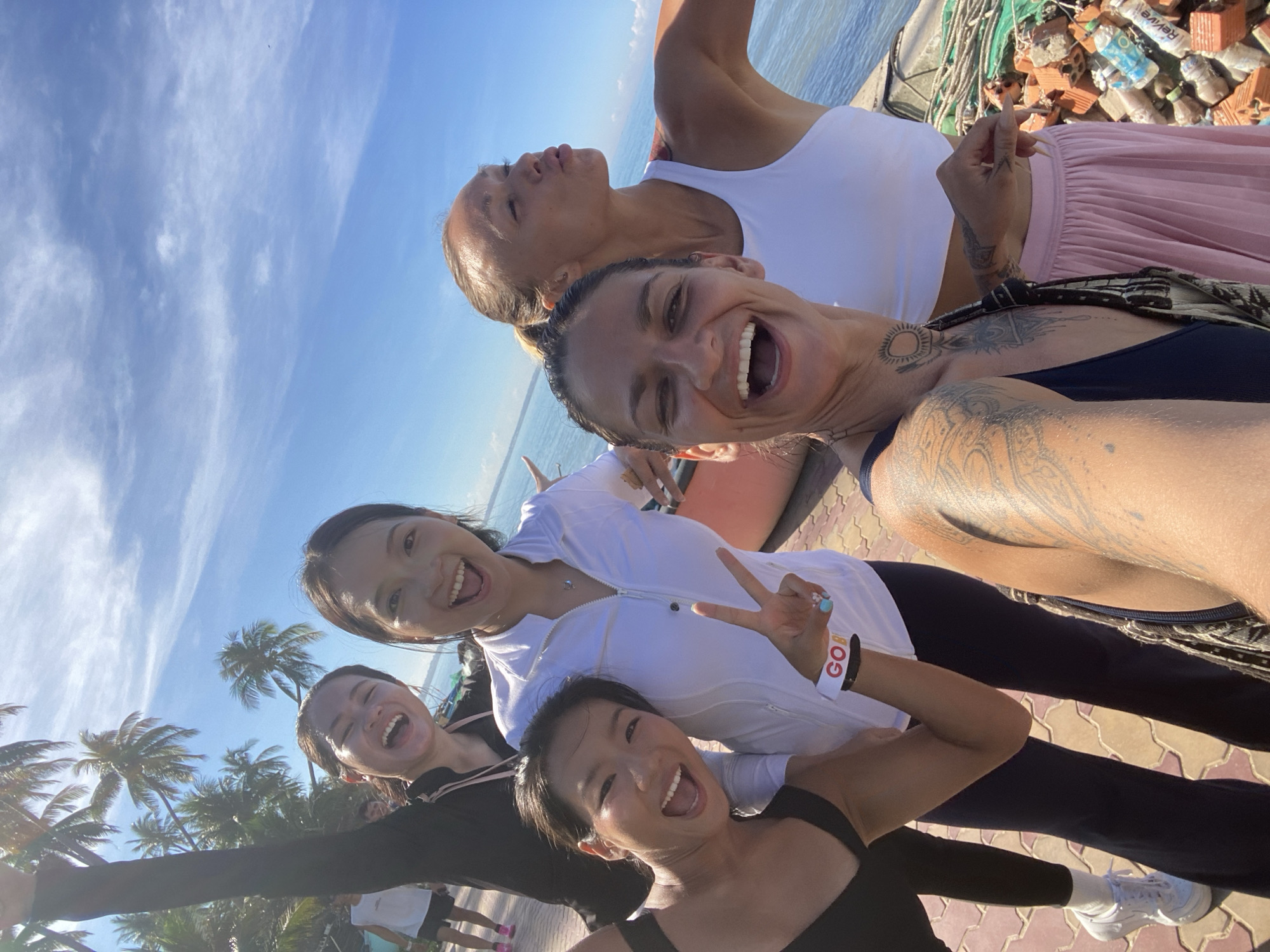 A group of five women are posing for a selfie in Mũi Né, Vietnam. The women are dressed in athletic wear and appear to be at a beach or coastal location, with palm trees and the ocean visible in the background. The sky is clear and blue, with some wispy clouds. The women are all smiling and appear to be in good spirits. Some of the women are making peace signs, while others are blowing kisses. The woman in the foreground on the right has tattoos on her arms and chest. There are also some piles of bricks and bottles visible in the background, suggesting a construction or cleanup site.