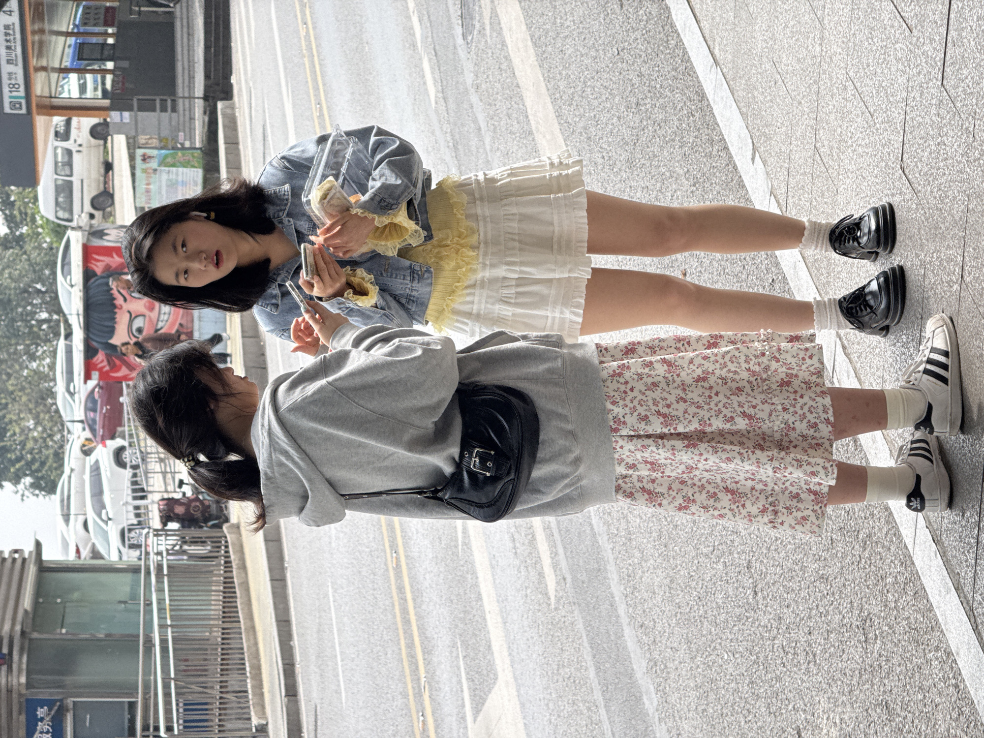 The image displays two young females standing on a grey tiled sidewalk adjacent to an asphalt road in Jiulongpo District, China, identifiable by a partially visible sign indicating "四川美术学院" (Sichuan Fine Arts Institute) and "18号".
The female on the left is observed from her left profile and back. She has dark hair styled in a ponytail with a black elastic. Her attire consists of a light grey hooded sweatshirt, a white skirt with a pink floral pattern, white socks, and white Adidas sneakers with black stripes. A black shoulder bag featuring a silver buckle closure is worn over her left shoulder. She is holding a smartphone with both hands, directing her gaze downwards towards the screen.
The female on the right faces towards the viewer with an open-mouthed expression. She possesses dark, shoulder-length hair and a light-colored earbud in her right ear. She is dressed in a light blue denim jacket over a yellow ruffled top, a white ruffled mini-skirt, white ruffled socks, and black chunky-soled shoes with laces. She holds two smartphones in her left hand and a clear plastic container containing food in her right hand.
The road behind them is marked with double yellow lines and exhibits patches of dampness. The background environment includes a metal railing, a glass structure, several parked vehicles (a white SUV, a red sedan, a white van), a large red and white mural depicting a cartoonish face, and additional urban structures, all under a clear sky.