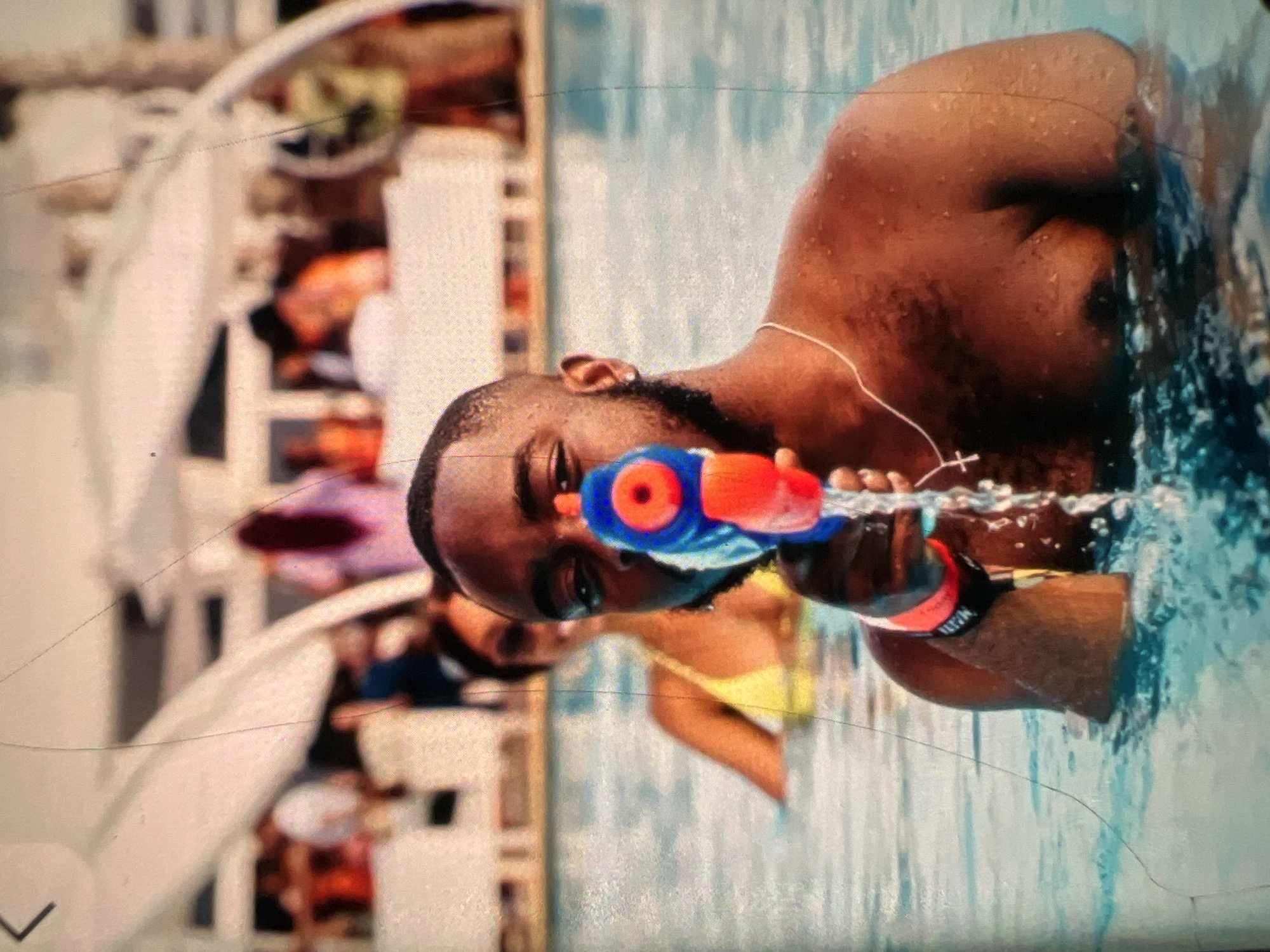 A Black man is in a swimming pool, holding a red and blue water gun and squirting water. He is wearing a silver necklace with a cross pendant and a black and red wristband. Behind him, a woman in a yellow bikini is also in the pool. In the background, there are other people and lounge chairs under white canopies, suggesting a resort or hotel setting. The water appears clear blue. The image quality is somewhat grainy, with some visible lines and scratches on the surface.