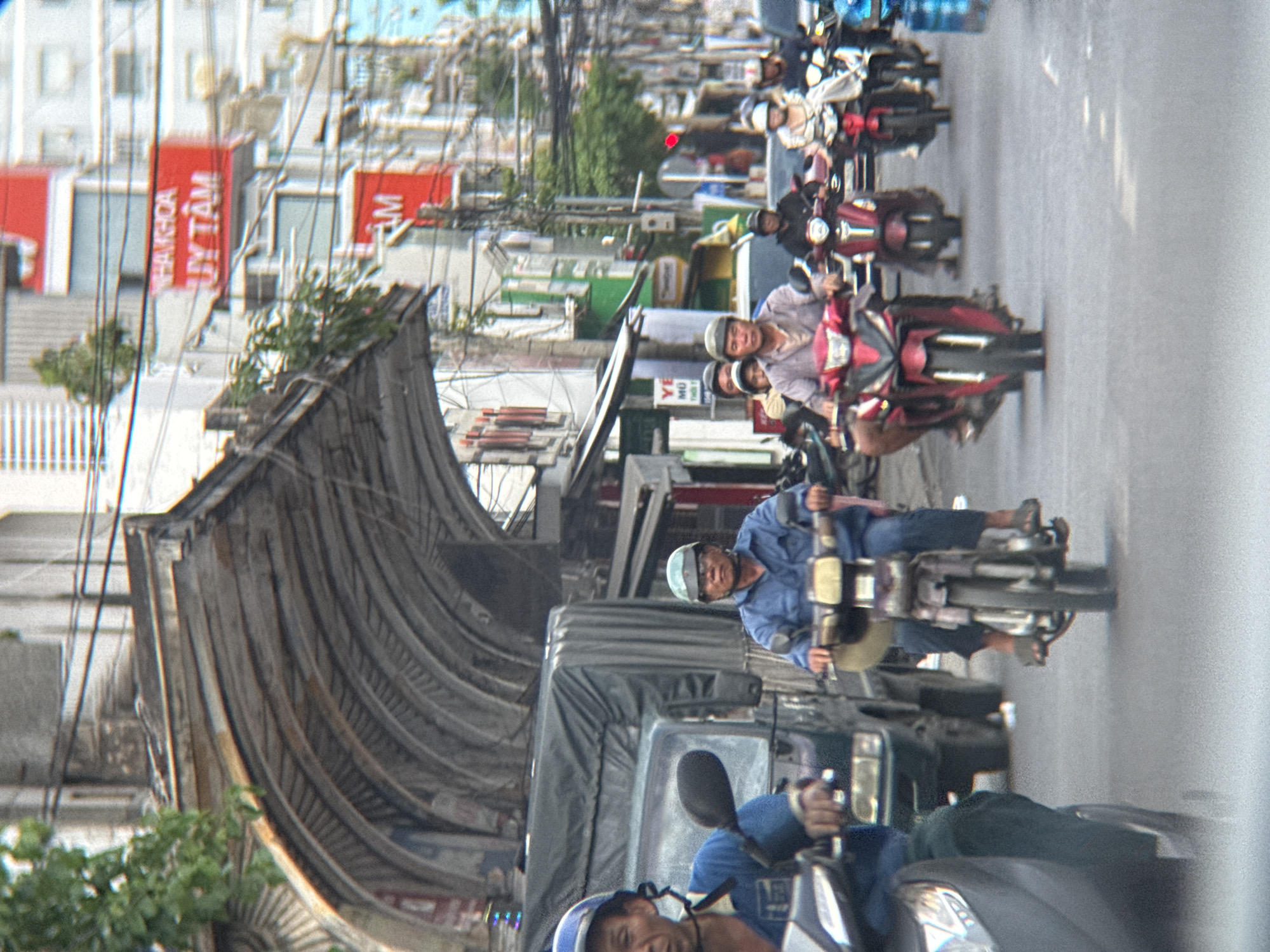 The image depicts a street scene in Nha Trang, Vietnam, likely during the daytime. Multiple individuals are riding motorcycles or scooters in what appears to be traffic. In the foreground, a person wearing a blue shirt and a blue helmet rides a scooter, partially visible on the left side of the frame. To their right, another person in a blue shirt and a blue helmet, wearing glasses, rides a scooter. Further down the street, several other motorcycles and riders are visible, some with helmets. The background shows buildings with signage, suggesting a commercial area. One sign in red reads "UY TAM". There are power lines and trees visible, contributing to the urban landscape. The road surface appears to be asphalt. The overall impression is one of everyday street life in a busy Vietnamese city.