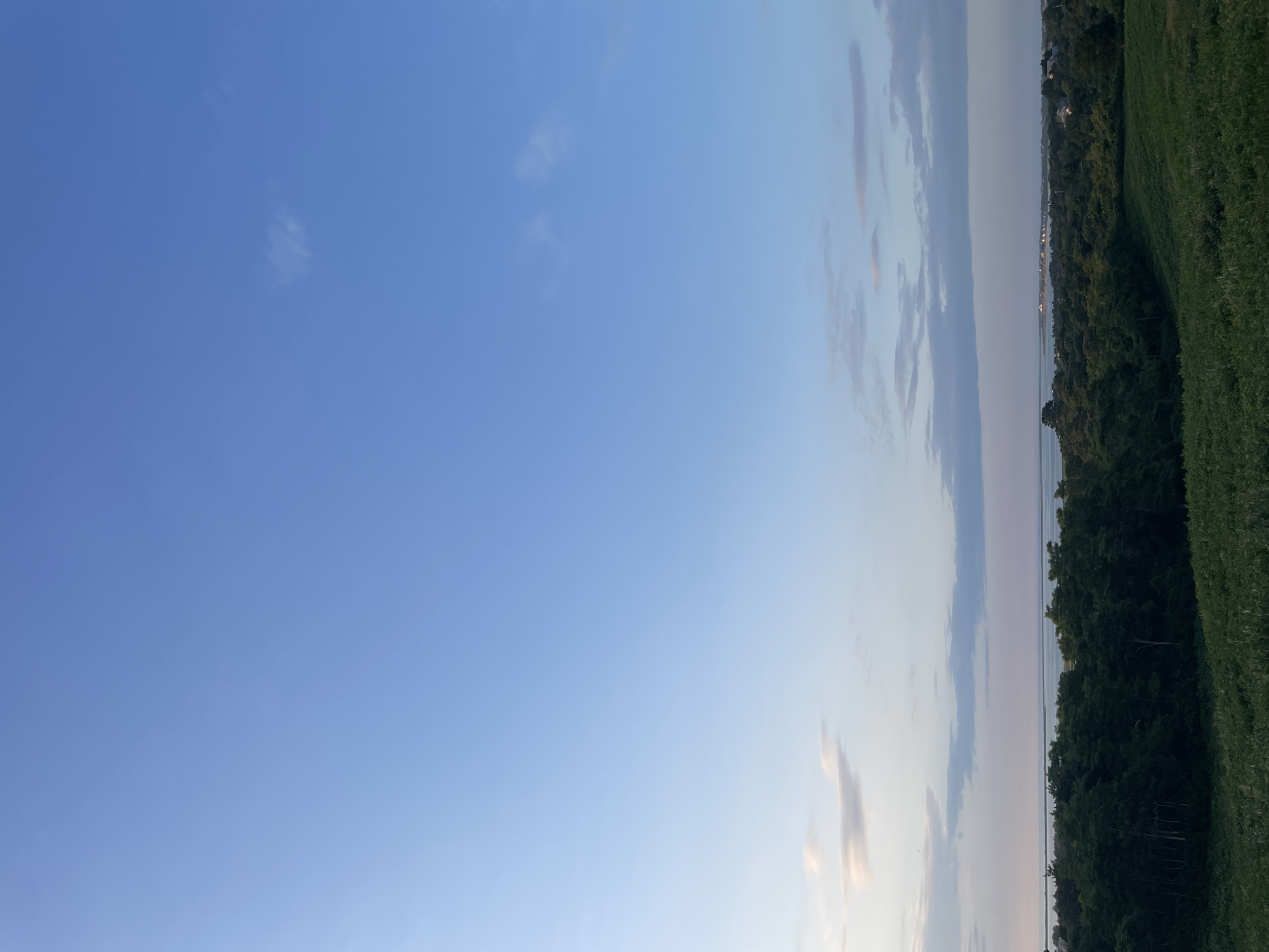 The image captures a serene landscape on the island of Nantucket, United States of America, during what appears to be late afternoon or early evening. The sky is a vast expanse of clear blue, transitioning to lighter shades near the horizon, with a few wispy, white clouds scattered throughout.
Below the sky, a body of water, likely the ocean or a bay, stretches across the mid-ground. The water's surface is calm and reflects the soft light of the sky. A distant coastline is visible, dotted with what seem to be houses and other structures, suggesting a settlement or town.
In the foreground and mid-ground, a dense canopy of lush green trees dominates the scene, creating a natural barrier between the viewer and the water. The trees are varied in height and texture, forming a rich, textured forest. In the very bottom of the frame, a field of green vegetation, possibly grass or low-lying plants, is visible. The overall impression is one of tranquility and natural beauty. There are no people or animals present in the image.