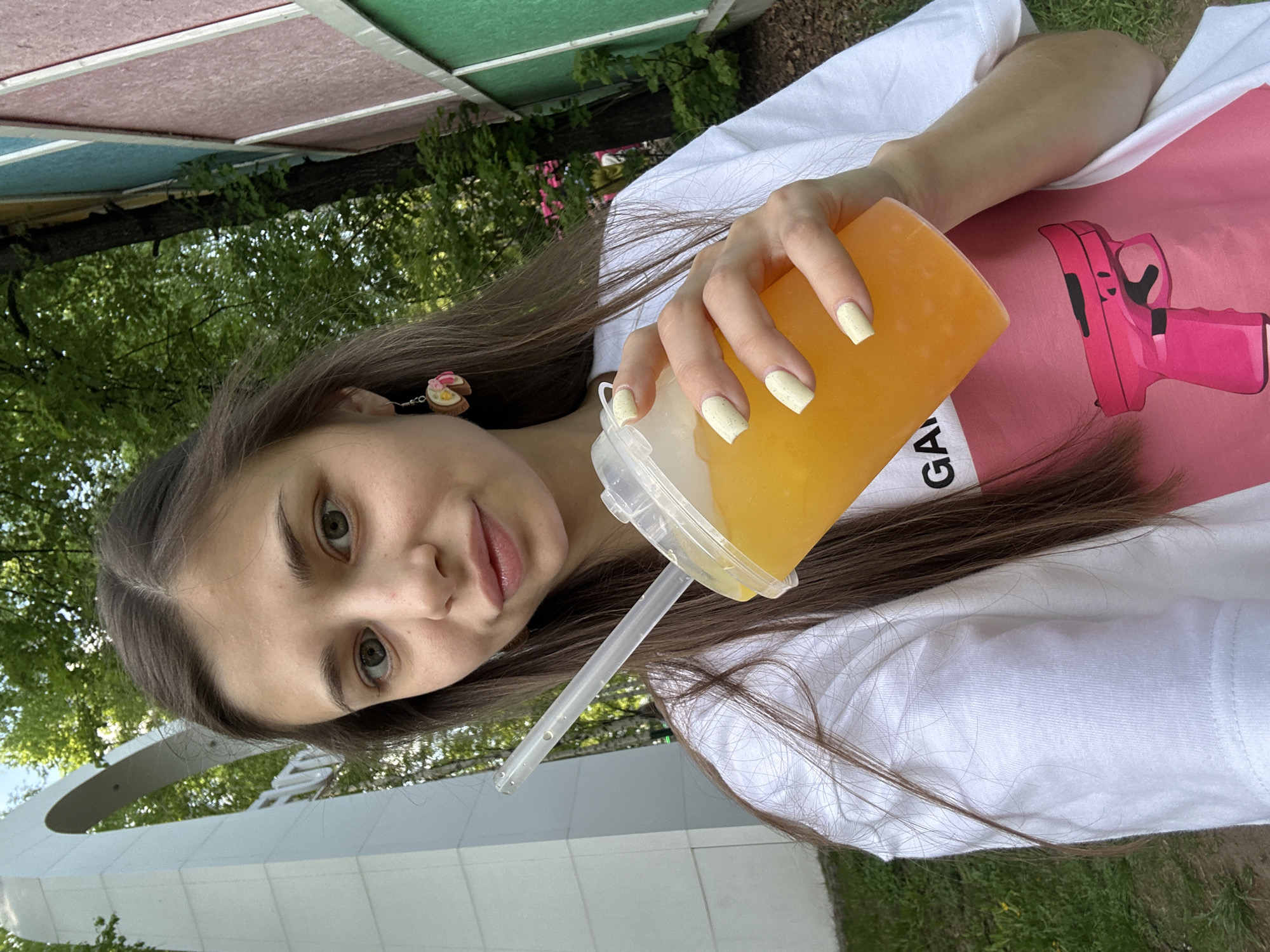 A young woman is holding a large cup of orange liquid with a straw, posing for a selfie or portrait. She has light brown hair and is wearing a white t-shirt with a pink graphic of a gun. She has light skin and is wearing pink lipstick and has light yellow nails. She is outside, with trees and greenery visible in the background. There are colorful buildings in the background.
Based on the structure in the lower-left corner of the image, the location is likely in Izhevsk, Russia, near the monument "Forever with Russia."
