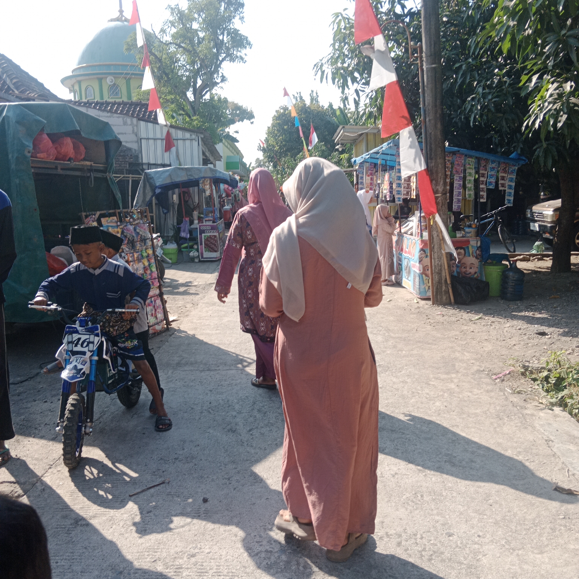 The image depicts a street scene in Tegowanu Kulon, Indonesia, with a mosque visible in the background. In the foreground, a young boy wearing a black peci cap and a blue jacket is sitting on a small dirt bike with the number "46" on it. He is looking forward, holding the handlebars. Behind him, two women are walking away from the camera, both wearing hijabs and modest clothing. The woman closer to the camera wears a peach-colored outer garment and a light-colored hijab, while the woman further away wears a maroon patterned outfit and a pink hijab. Several stalls are set up along the side of the road, selling various items, including what appear to be packaged snacks and drinks. Red and white Indonesian flags are visible, adding a festive touch. The ground is paved with concrete, and shadows suggest a bright, sunny day. Trees and other vegetation line the street. A silver vehicle is parked on the right side of the road.