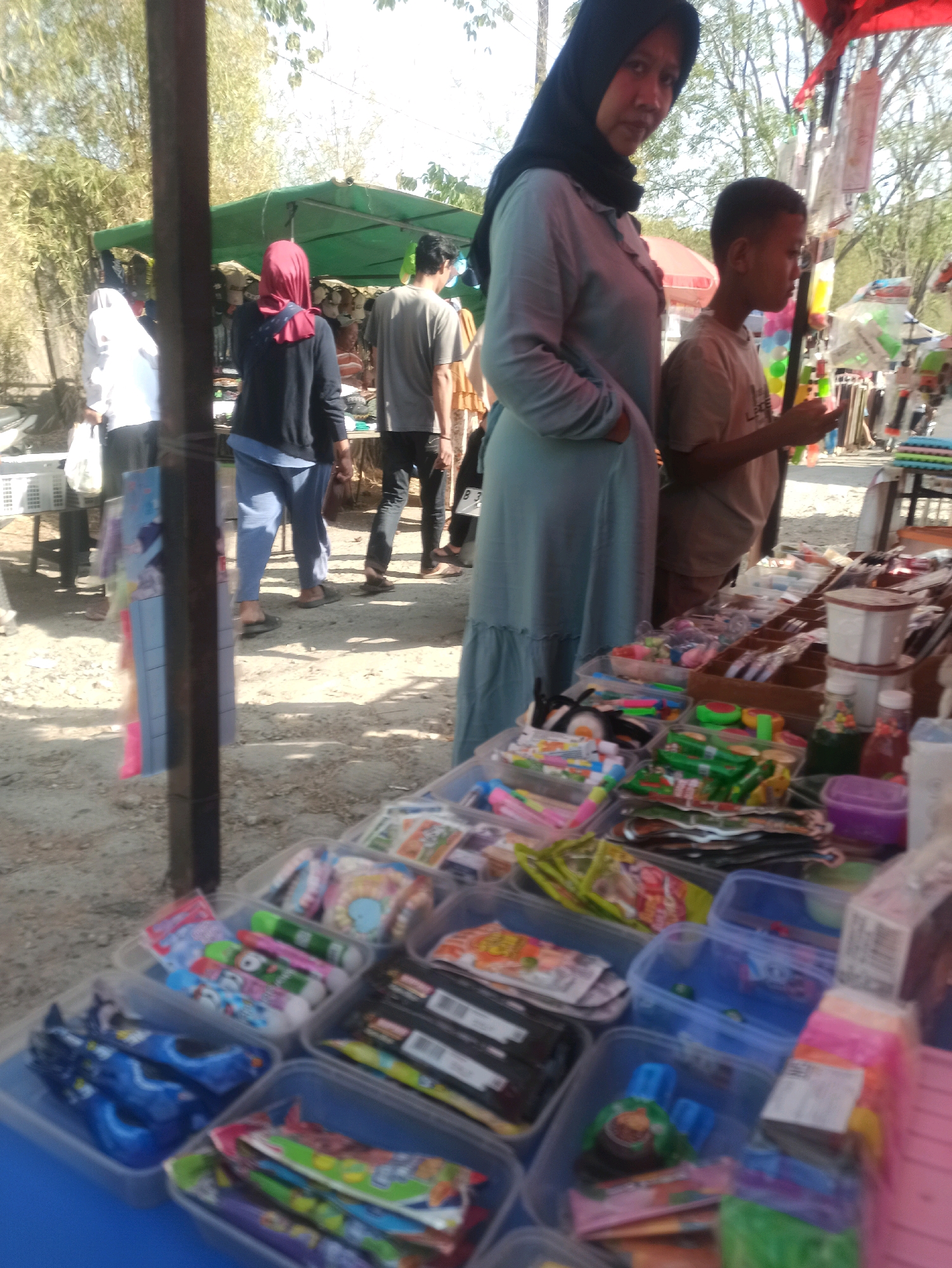 An outdoor marketplace is shown in Telagamurni, Indonesia. In the foreground, several plastic containers filled with various items, likely school supplies or small toys, are arranged on a blue table. These items include colorful pens, pencils, erasers, and packaged snacks. A woman wearing a black hijab and a light blue dress stands to the left of a young boy, who is wearing a light brown t-shirt. They are looking towards the items on display. In the background, other people can be seen browsing at market stalls under green awnings. The ground is covered with sand and dirt. The overall scene suggests a typical local market environment.