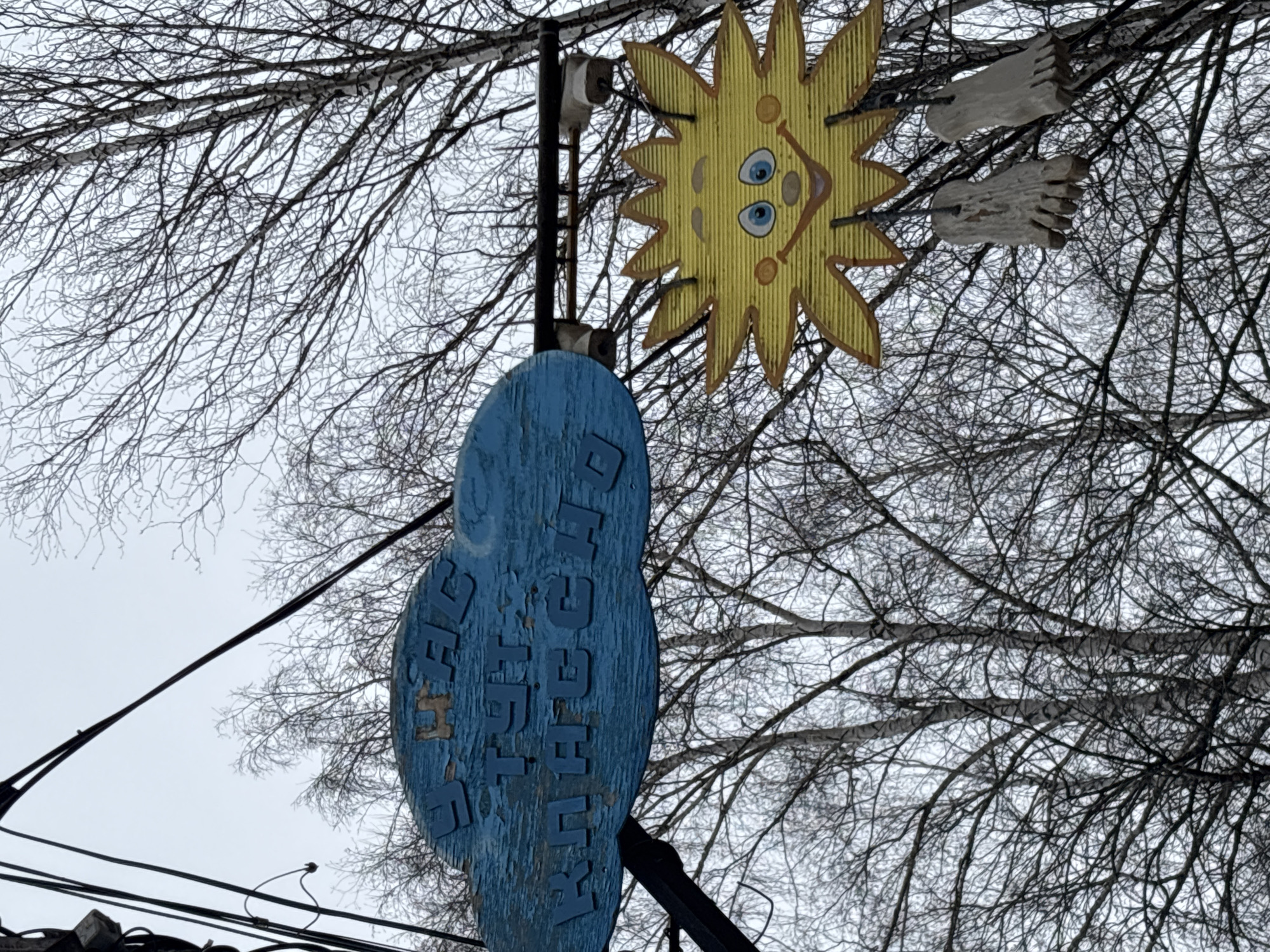 In the city of Chaikovsky, Russia, a street sign consisting of a sun with a cartoon face, a pair of cartoon feet, and a blue oval sign with Cyrillic letters is attached to a metal pole. The sign is situated against a backdrop of bare tree branches and an overcast sky. Power lines run near the base of the sign. The blue oval sign shows signs of age and wear, with peeling paint.