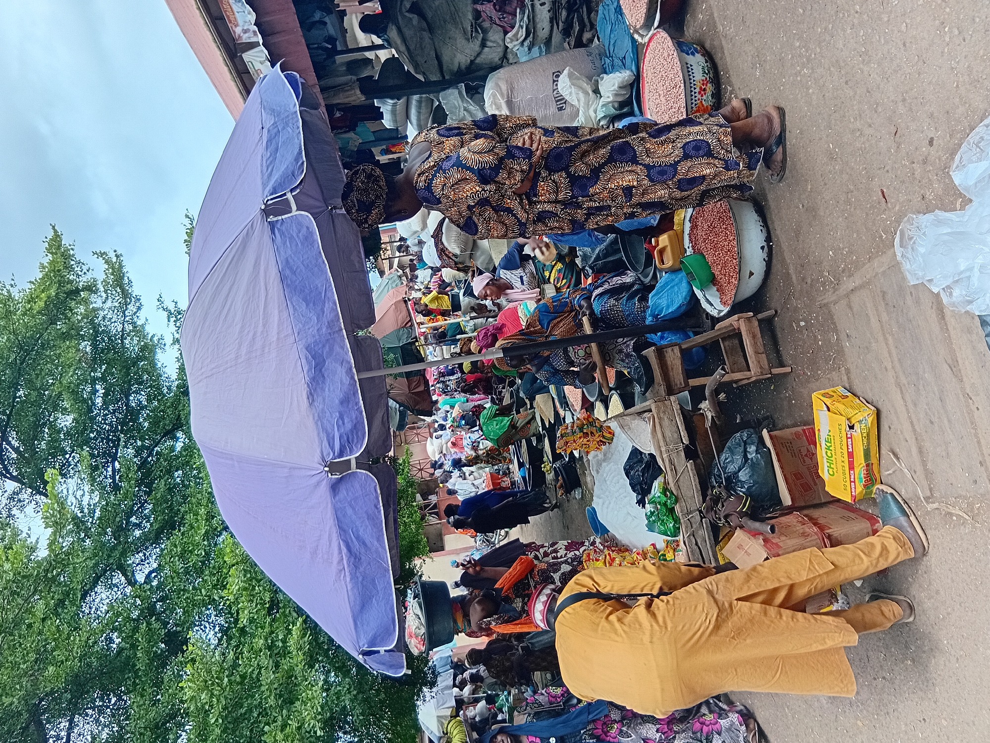 This is a forensic, matter-of-fact description of a street market scene in Lokoja, Nigeria. The image captures a bustling marketplace under a large, purple umbrella, with numerous individuals engaged in commerce. In the foreground, a person dressed in a yellow garment is bending over, seemingly attending to goods on display. To their right, a woman in a vibrant, patterned blue and gold dress stands with her back to the camera, also facing the market stalls.
The market appears to be selling various food items, prominently displayed in bowls and sacks. Large quantities of red beans are visible in white bowls, suggesting a food market. Behind the woman in the patterned dress, sacks of grain or other produce are stacked high. Several other people, dressed in colorful traditional clothing, are scattered throughout the scene, some appearing to be vendors and others customers. The environment is an outdoor setting, with trees visible in the background and a building with a corrugated roof to the right. The ground is paved with concrete. The overall impression is one of an active and vibrant local market.
