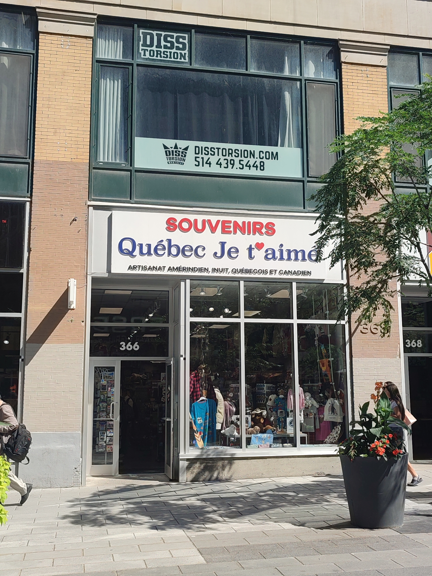 This image captures a street-level view of a store in Montreal, Canada, located at 366 and 368 on a street. The store's facade features large display windows and a prominent sign that reads "SOUVENIRS Québec Je t'aime" with the tagline "ARTISANAT AMÉRINDIEN, INUIT, QUÉBÉCOIS ET CANADIEN." Above this, on the second floor, another sign displays "DISS TORSION" and a website address. The store's interior is visible through the windows, showcasing various merchandise, including clothing and souvenirs.
Two individuals are present. A person on the left walks away from the store, carrying a black backpack. On the right, two women walk past the store. One is carrying a light pink shopping bag. A large dark planter with orange flowers and green foliage is positioned on the sidewalk in front of the store. The building's exterior is a mix of brick and concrete. The sidewalk is paved with interlocking stones, and the dappled shadow of a tree falls across it. The weather appears to be sunny.