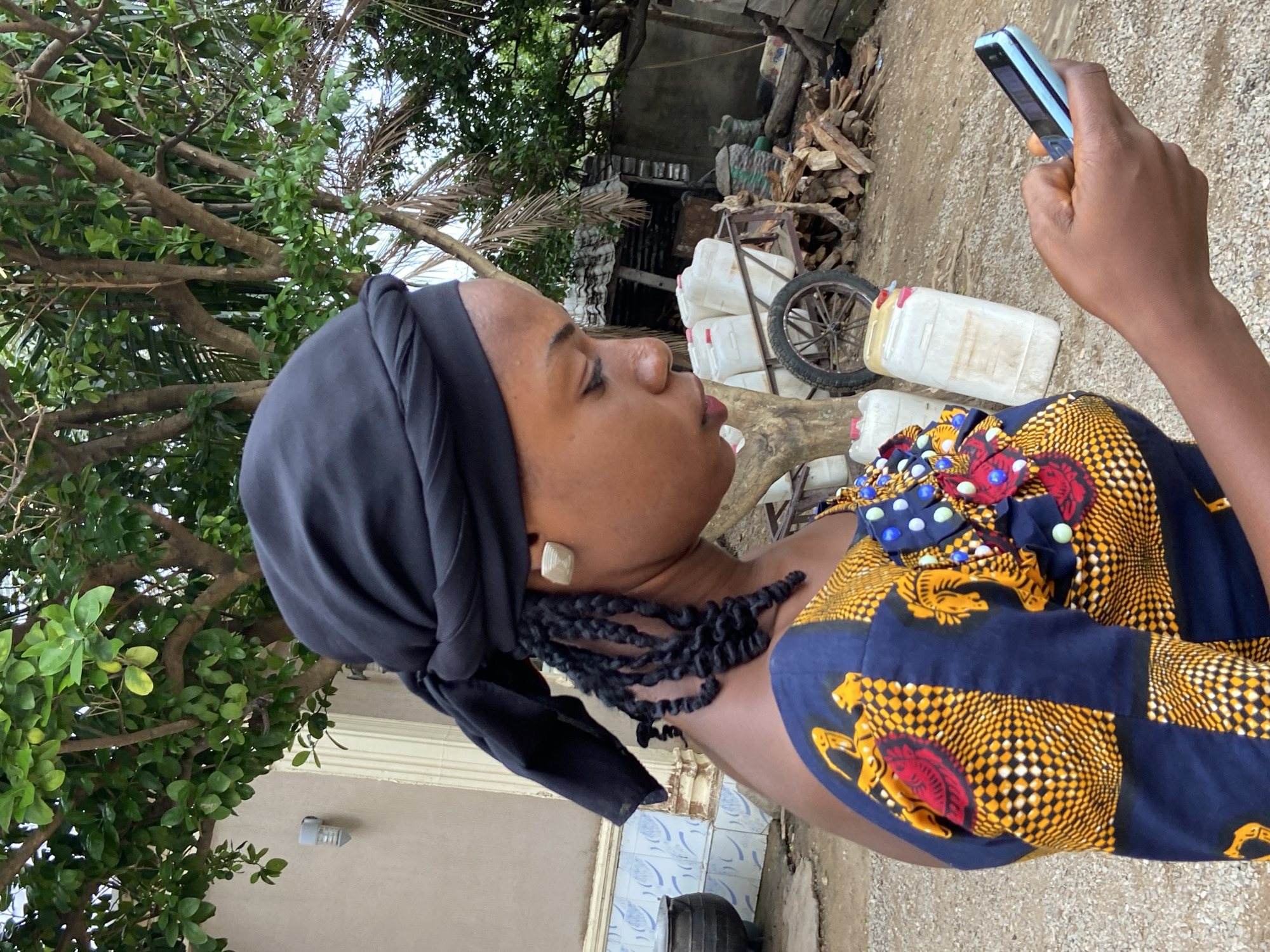 A woman wearing a dark head wrap and a colorful, patterned dress with decorative accents is holding a blue phone. She is outdoors, likely in a yard or near a building. In the background, there's foliage, firewood, stacked containers, and a wheel, suggesting a semi-rural environment in Mararaba, Nigeria. The woman seems to be focused on her phone.