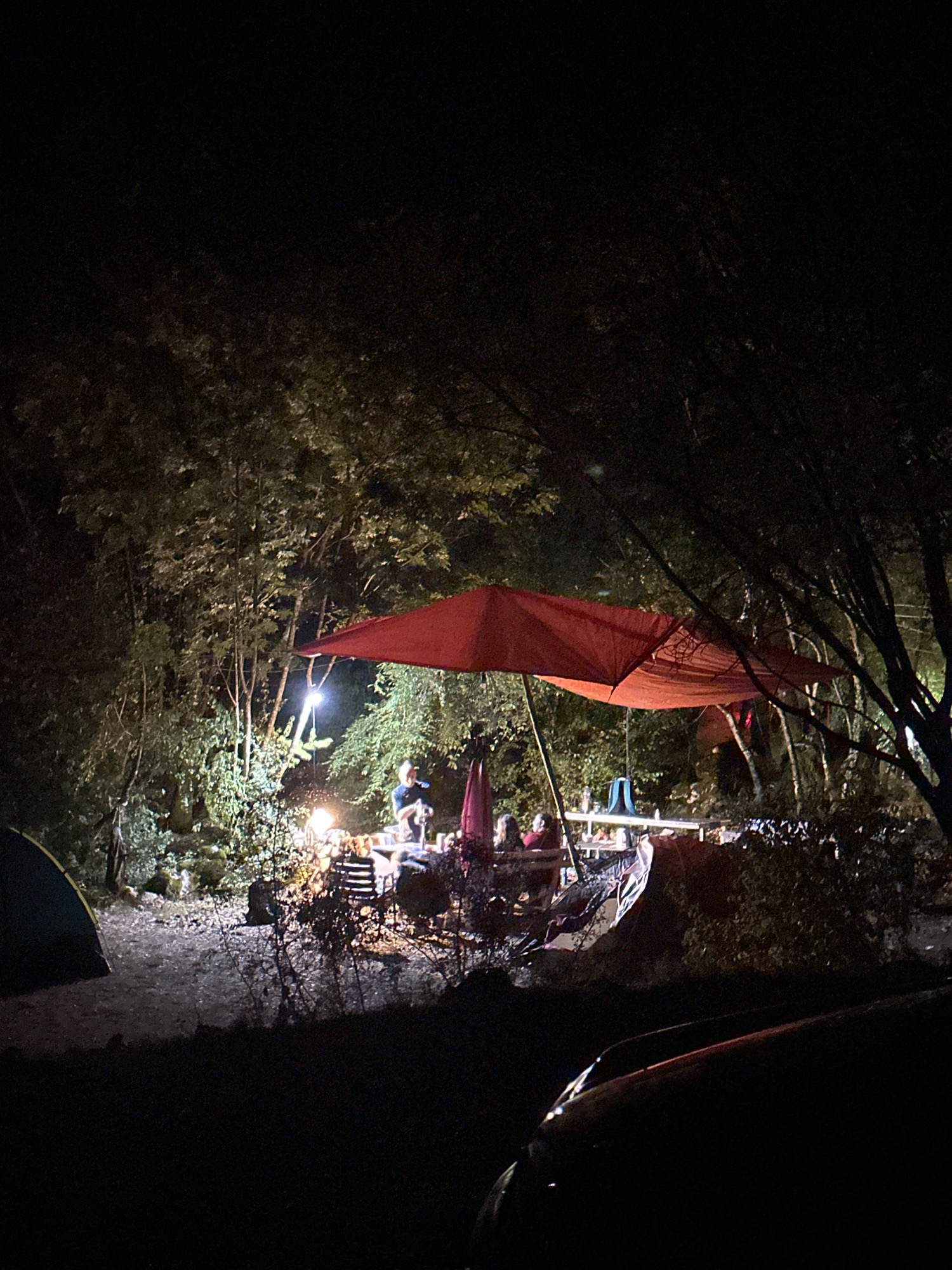 The image depicts a campsite at night, likely in a wooded area, under a dark sky. A bright red umbrella is prominently featured, providing shelter over a picnic table where several people are seated. A campfire is visible, casting a warm glow on the surroundings and illuminating the faces of those gathered around it. A tent is pitched to the left of the frame, indicating overnight accommodation. The foreground is dominated by the dark silhouette of a car. The environment appears to be natural, with dense foliage and trees encircling the campsite. There is no indication of the specific location within Cetinje, Montenegro, beyond it being a nocturnal outdoor scene.