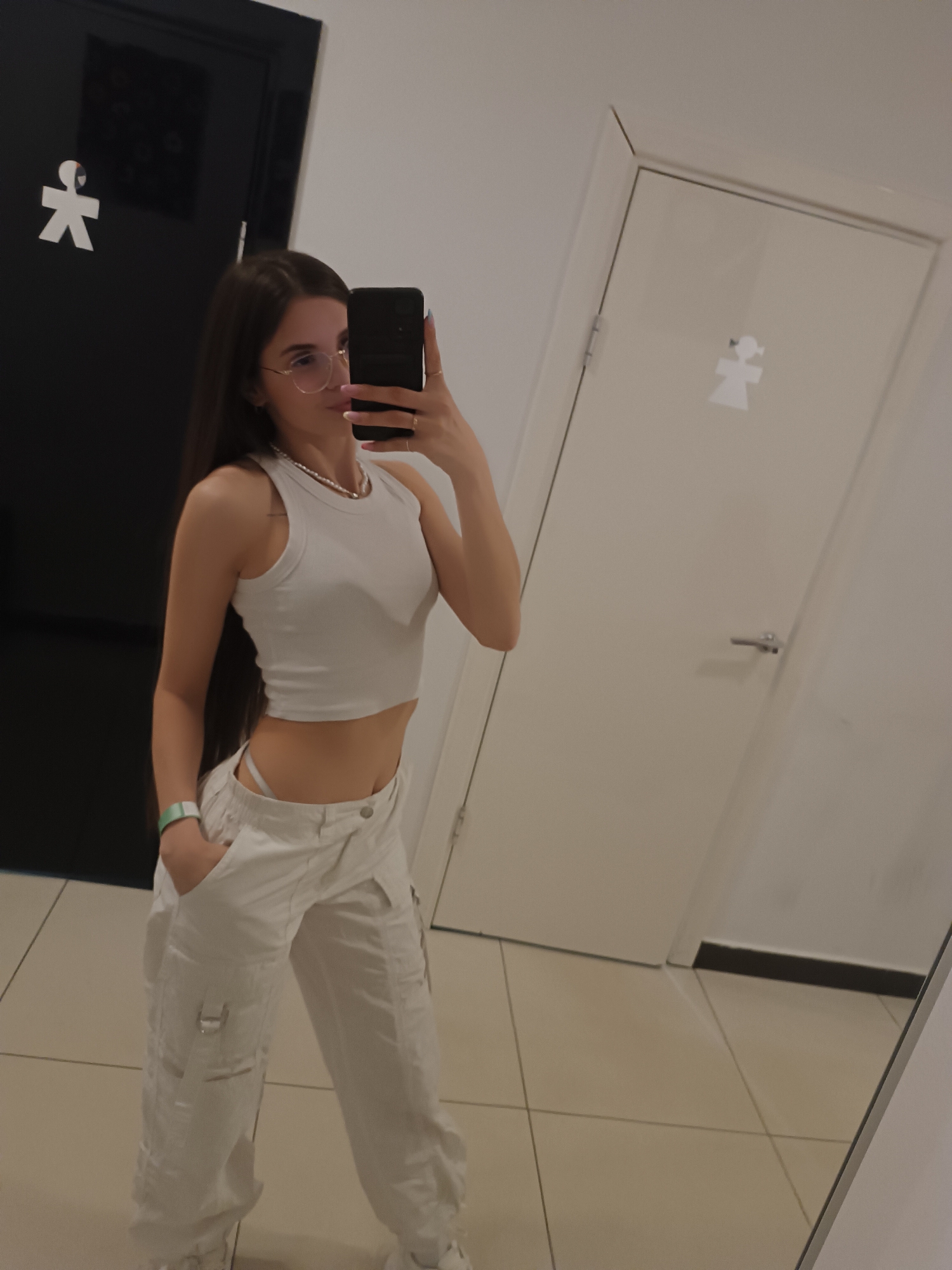A young woman stands in front of a mirror, taking a selfie. She wears a white sleeveless top and white cargo pants. The location appears to be a public restroom, indicated by the gender symbols on the doors behind her. The restroom has a tile floor and white or light-colored walls. The location is likely not in Chaikovsky, Russia, as restroom signs with these symbols are not exclusive to that city.