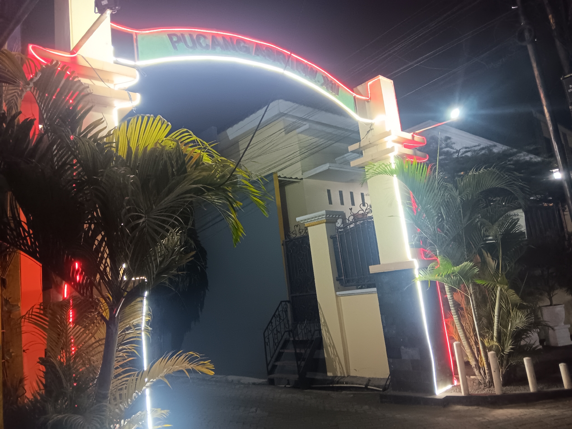 The image shows an illuminated archway at night in Batursari, Indonesia, with neon lights forming the words "PUGANG ASRI XV". The archway is adorned with red and white neon lights. Tropical palm trees are visible in the foreground, their fronds illuminated by the lights. Behind the archway, a building with a pale yellow facade and a white roof is visible. The building has dark windows and a decorative metal gate. A set of outdoor stairs leads up to the gate. Electrical wires are strung overhead. The ground appears to be paved with bricks or cobblestones. The overall scene suggests an entrance to a residential complex or a business establishment.