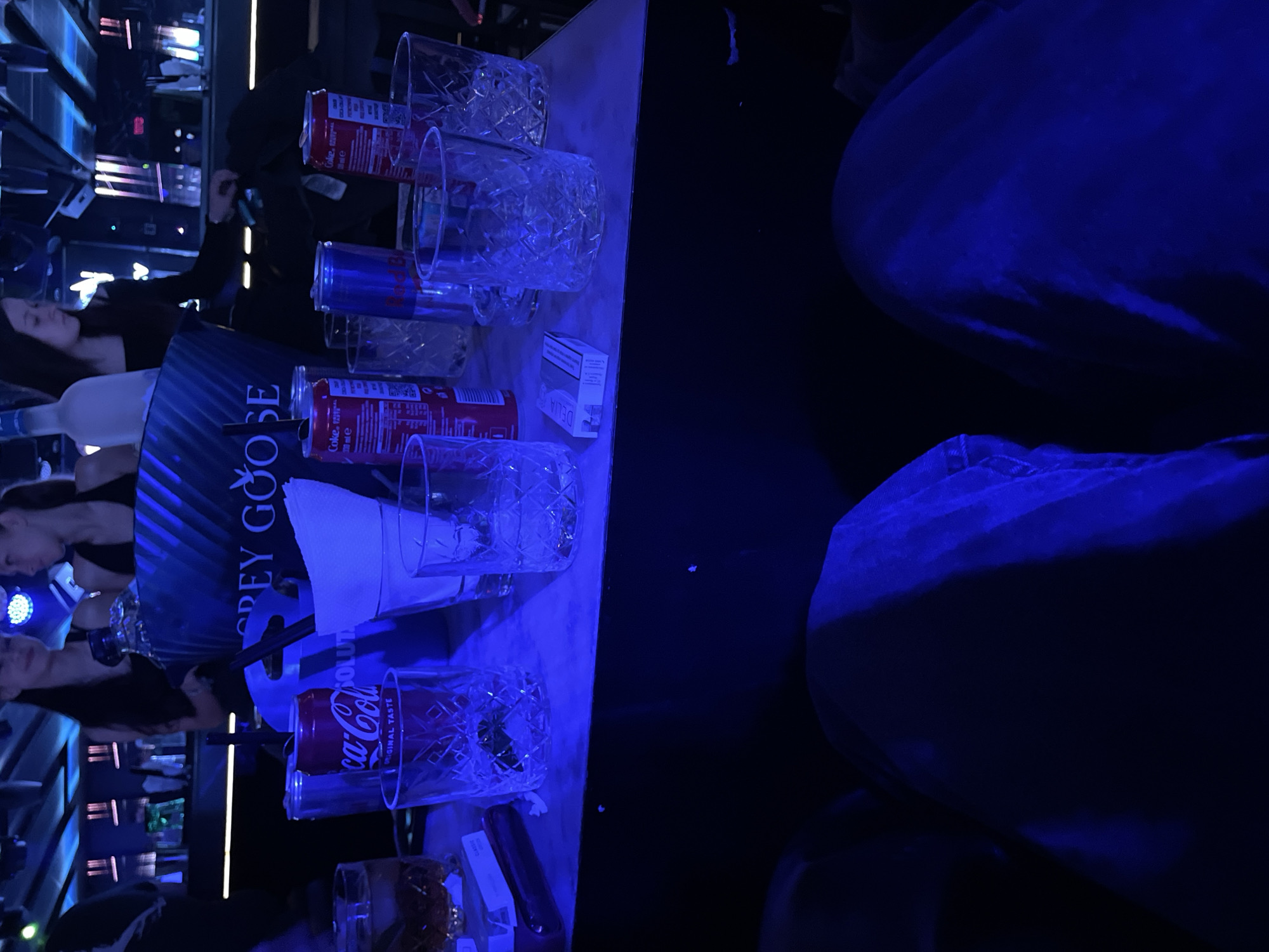 The image depicts a dimly lit bar or nightclub setting, likely in Sofia, Bulgaria. Multiple patterned glasses are arranged on a counter, some containing Coca-Cola and Red Bull cans. A bottle of Grey Goose vodka is present, accompanied by straws and white napkins. A packet of "Delia" brand cigarettes also sits on the counter. Partial figures of individuals, including people, are seen in the background of the image.