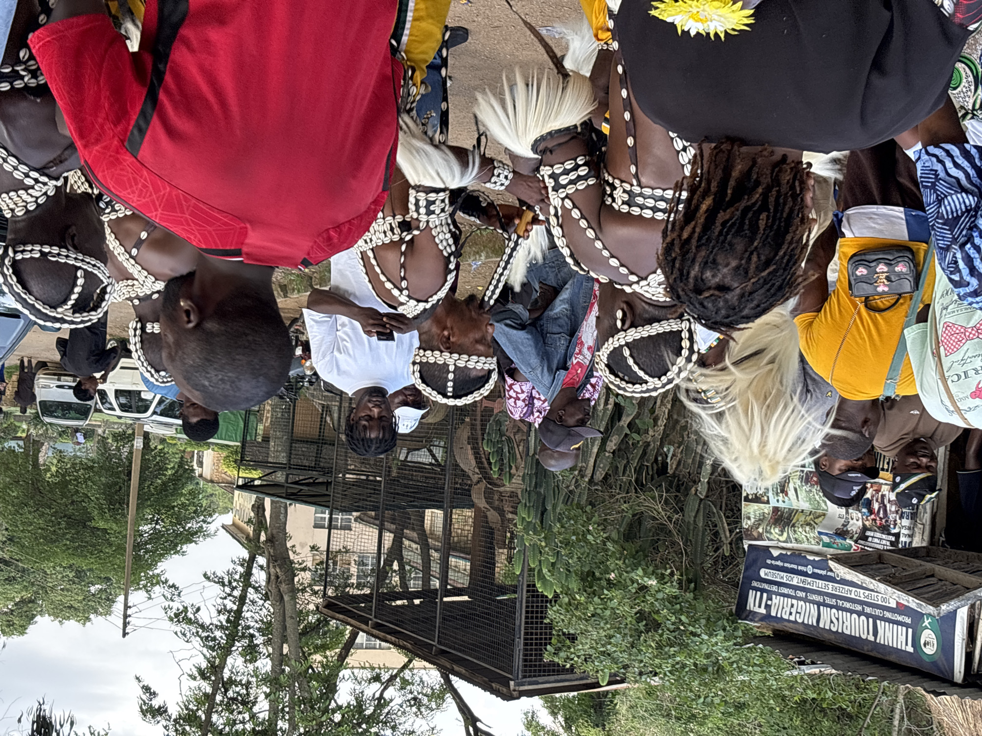 This image captures a scene in Jos, Nigeria, likely at or near the National Museum Jos, which is known for its cultural artifacts and heritage. In the foreground, several individuals are gathered, some dressed in traditional attire adorned with cowrie shells and white tassels, suggesting a cultural performance or ceremony. One man in the center is playing a musical instrument, possibly a flute, while others observe. Behind them, a banner for "THINK TOURISM NIGERIA-TTN" is visible, promoting cultural and historical sites. The environment includes trees, a building with caged enclosures, and parked vehicles, indicative of an outdoor public space or institution. The cloudy sky suggests it might be an overcast day. The people appear to be a mix of performers and spectators, with a sense of community and cultural engagement.