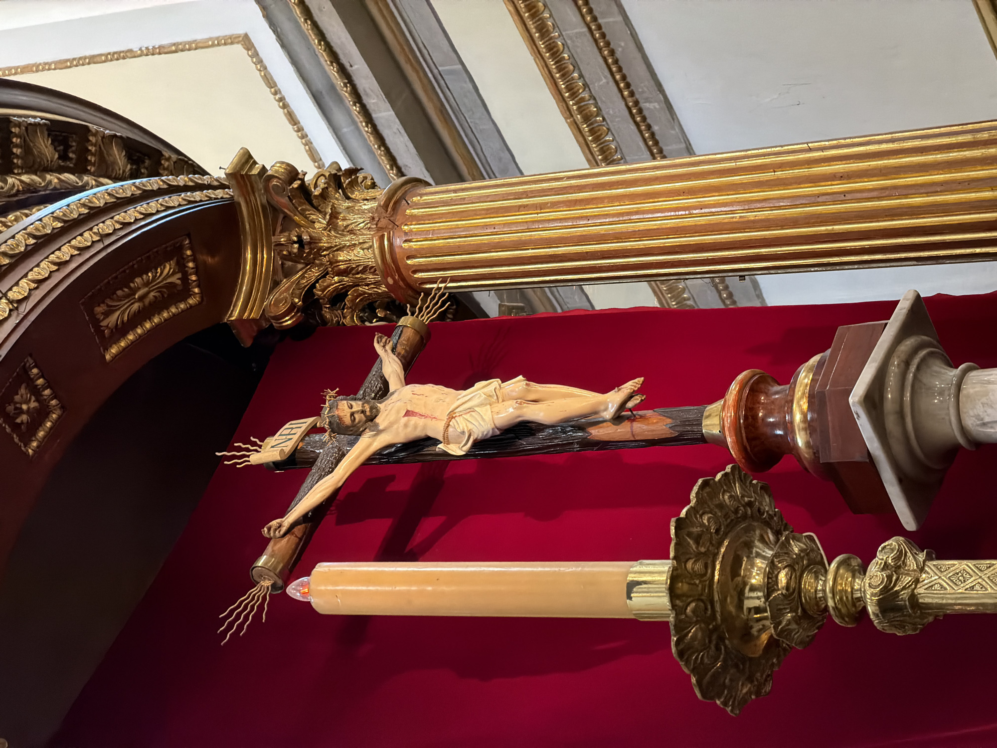The image captures a view inside a church, likely in Puebla, Mexico, evidenced by the architectural details and religious iconography. The focal point is a crucifix with a depiction of Jesus Christ, mounted on a cross. A candle holder with a tall candle is visible. The background is a deep red, possibly a wall or drape. Ornate gold-leafed architectural elements, including columns and architraves, indicate a baroque or similar style, common in Mexican churches of the colonial period. The scene evokes a sense of religious reverence and historical artistic craftsmanship.