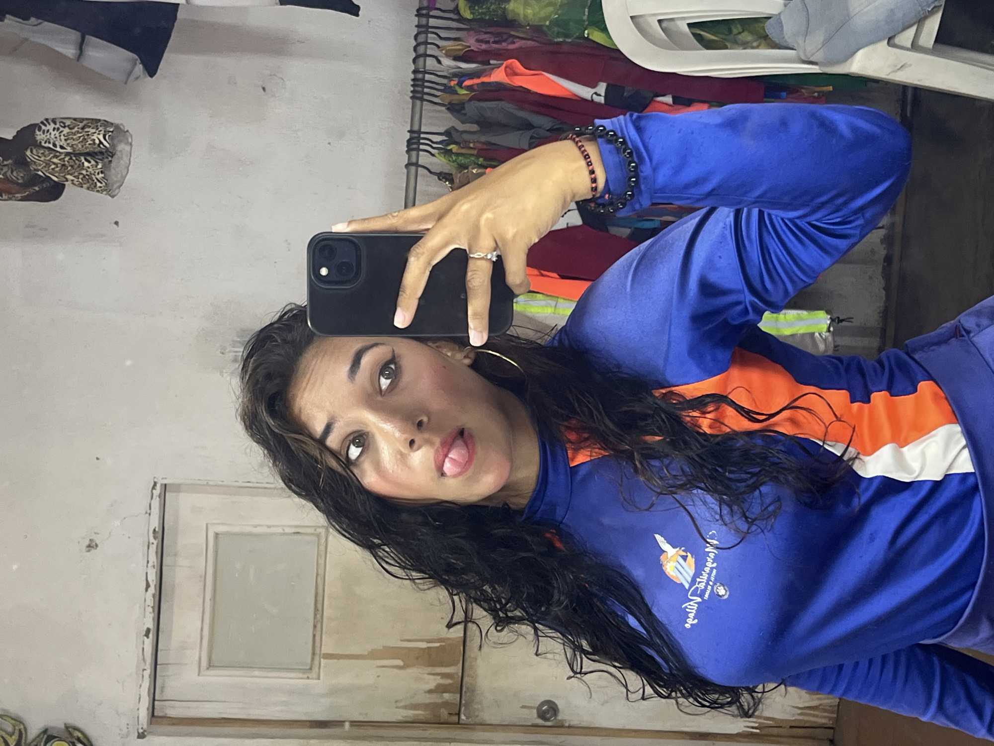 A woman is taking a selfie in what appears to be an indoor setting. She is wearing a blue and orange athletic jacket with a logo and text on the chest. The woman has long, dark, wavy hair and is wearing hoop earrings. Her tongue is slightly extended. She is holding a black smartphone in her left hand. On her left wrist are two bracelets. She is wearing a ring on her ring finger.
In the background, there is a rack of clothes. Additionally, there's a white door and wall.
It is difficult to confirm the location as Porlamar, Venezuela without further contextual clues.