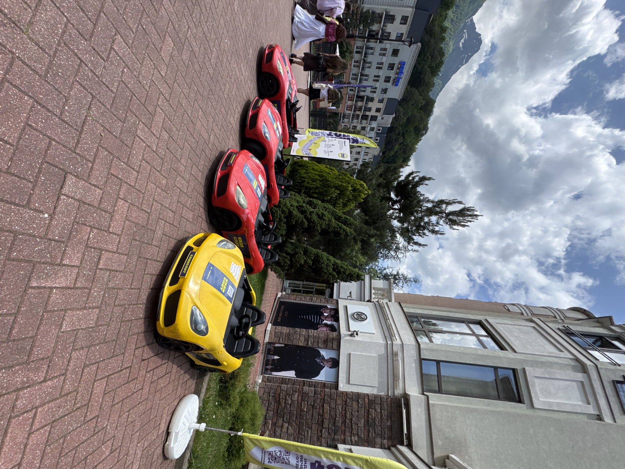 The image captures a bright outdoor scene in Estosadok, Russia, likely on a summer day given the clear sky with scattered clouds and lush greenery. In the foreground, a row of four colorful toy electric cars is parked on a red brick-paved pathway. Three are bright red, and one is yellow. They are all low-profile, sports-car-style vehicles with black interiors and wheels. Each car has some visible branding and decals, with the yellow car featuring a blue and yellow stripe across its hood and the text "КИД СПОРТ" and a web address.
Behind the toy cars, a modern, light-colored building with a stone-clad lower section dominates the right side of the frame. Large windows and architectural details are visible on its facade. On the building's exterior, there are two large posters displaying fashion models, with branding that includes "PAUL&SHARK".
To the left of the building, a multi-story hotel with a blue sign reading "Park Inn" is visible, nestled against a backdrop of green trees and a majestic snow-capped mountain in the distance. Several people are walking on the pathway to the left of the toy cars, including two women in dresses and two women in casual attire. One woman is wearing a long white dress, another a purple top and dark skirt, and the other two are wearing dark outfits.
A tall, yellow flag with text and a QR code stands prominently to the right of the toy cars, near the building. Various green trees and bushes are scattered throughout the scene, adding to the natural beauty of the mountainous resort town. The overall impression is one of a recreational area within a resort, likely catering to families and offering activities for children.