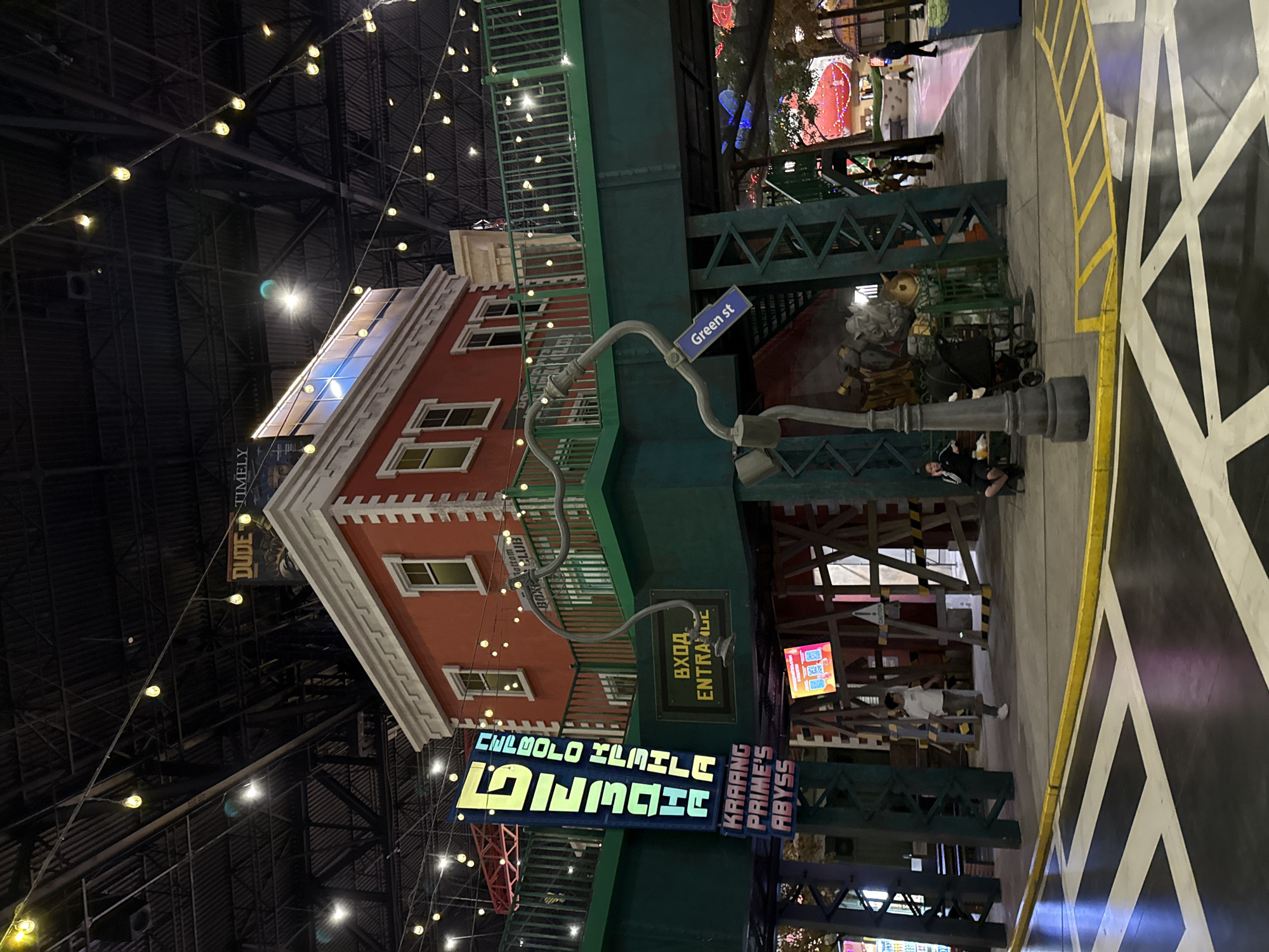 This image captures an indoor entertainment venue in Moscow, Russia, designed to resemble an urban streetscape. Central to the left is a two-story reddish-brown building facade with white trim and multiple windows, reminiscent of a city brownstone. Above and in front of it, a dark green elevated structure, possibly an overhead train track or bridge, dominates the midground, supported by metal beams.
The background reveals a vast, dark ceiling with exposed metalwork and numerous small, yellow string lights, creating an artificial nighttime atmosphere. The ground features a bold black and white geometric pattern with yellow markings, simulating a patterned street or plaza.
Several people are visible in the scene: one person is seated near a grey, industrial-style street lamp, while another in a white shirt stands on the patterned floor. Other individuals can be seen walking in the background.
Notable signage includes a blue "Green st" sign, a yellow sign indicating "ВХОД ENTRANCE" (Russian and English for "Entrance"), and a large, colorful, neon-style sign that prominently displays "KRAANG PRIME'S ABYSS" alongside stylized, less legible text that appears to read "CHEWBOLO HMILE NINJACH". A faint banner in the far background shows "DUDE TIMELY". The presence of Russian text confirms the location context. The scene conveys a relaxed, recreational ambiance within a themed environment.
