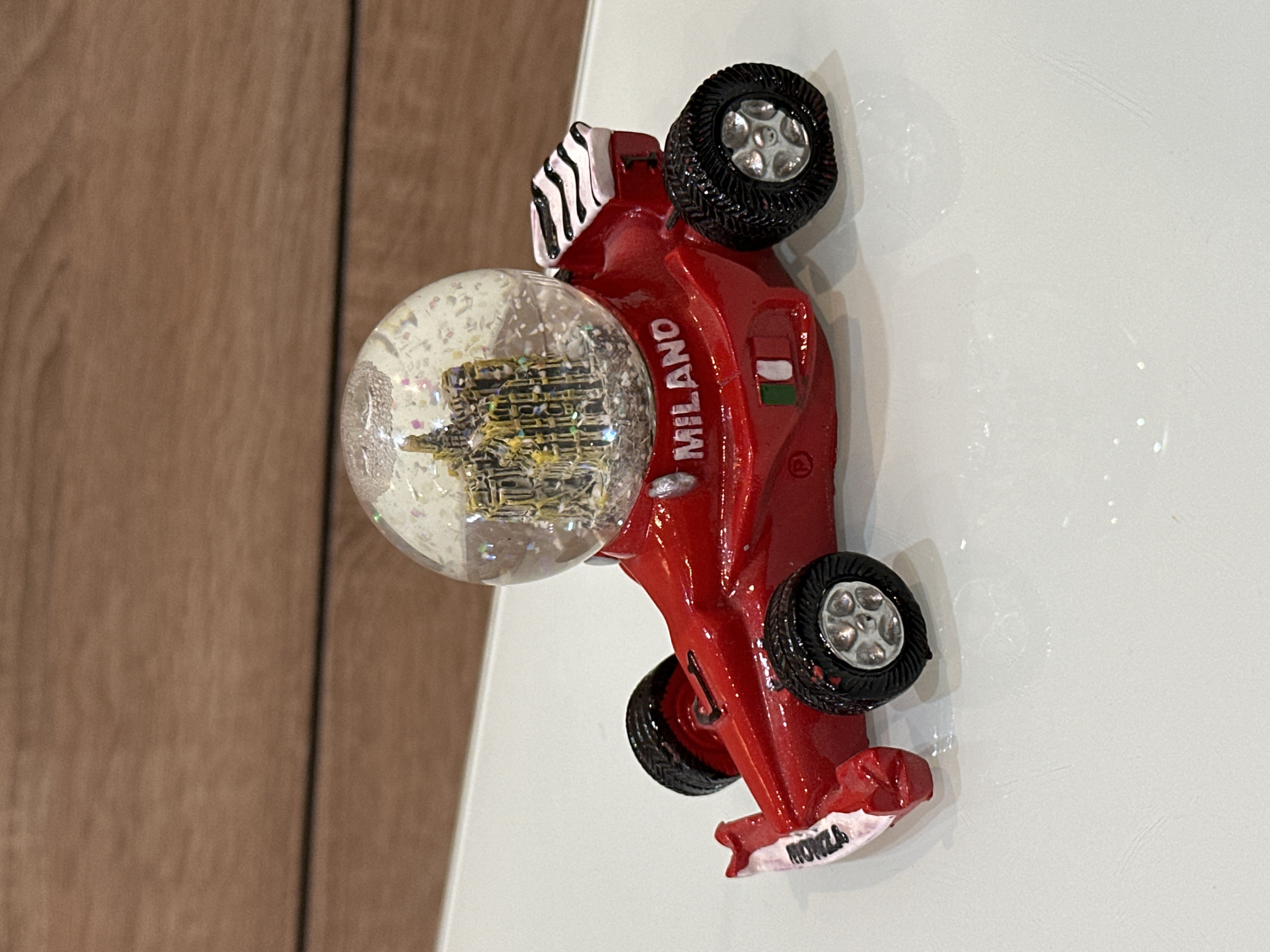 The image shows a red toy race car with the words "MILANO" and "MONZA" on it. The car has an Italian flag on its side and a snow globe attached. The location is not specified in the image.