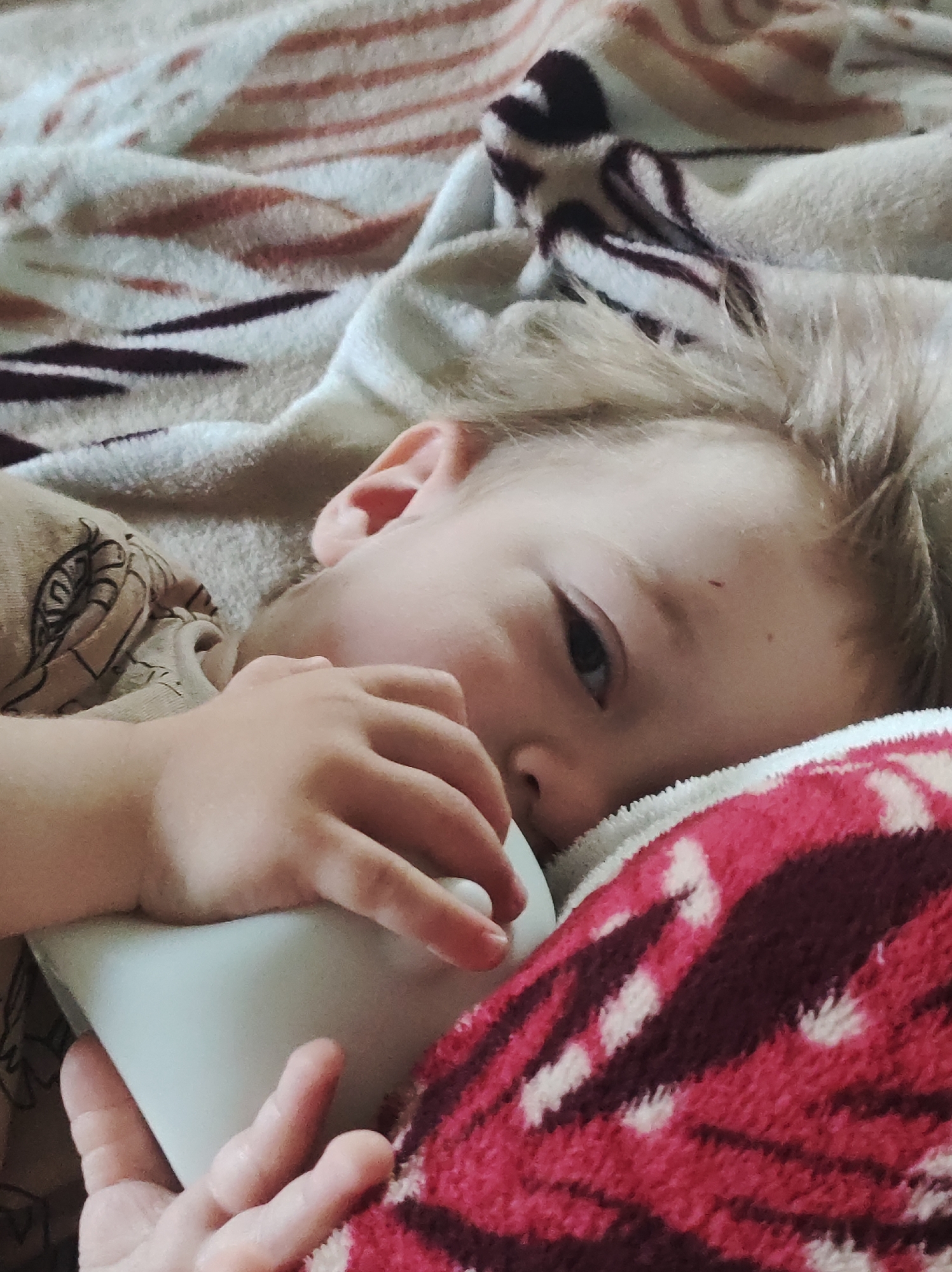 A young child is lying on a bed, holding a white bottle in their hands. They are partially covered by a blanket with a pink and brown pattern. The child's head rests on a pillow with a similar pattern. The location appears to be indoors, likely a home, but the exact city and region are unknown.