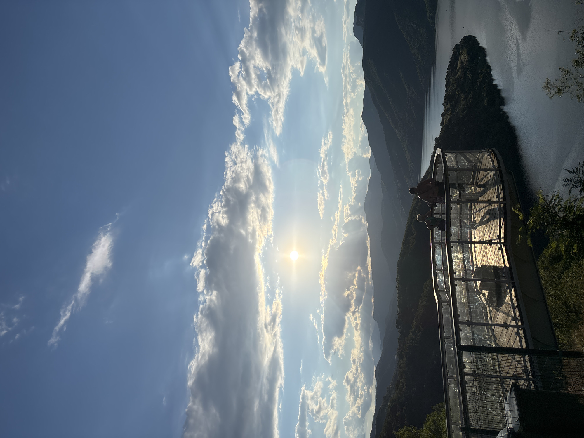This is a vertical, wide-angle, eye-level shot of a scenic overlook in the city of Borovitsa, Bulgaria, capturing the dramatic contrast between the sky and the landscape during a late afternoon sunset. The upper portion of the image is dominated by a vast expanse of blue sky dotted with a variety of clouds, ranging from wispy cirrus formations to larger, more dense cumulus clouds that are illuminated by the setting sun. The sun itself, a bright, intense orb, is partially obscured by a bank of clouds, casting rays of light that pierce through the atmosphere, creating a luminous, ethereal effect.
Below the sky, a series of layered mountain ranges recede into the distance, their peaks silhouetted against the bright sky and bathed in the warm glow of the sunset. The mountains appear lush and covered in dense vegetation, their slopes fading into a hazy, atmospheric perspective. A large body of water, likely a lake or reservoir, is visible in the mid-ground, its surface reflecting the subtle colors of the sky and the surrounding landscape. The water's edge is bordered by a peninsula covered in trees.
In the foreground, a modern observation deck juts out over the landscape, providing a vantage point for visitors to admire the view. The deck features a glass railing that offers unobstructed sightlines, and its wooden floor is illuminated by the low-lying sun, creating long, dramatic shadows. Two individuals are present on the deck, silhouetted against the bright backdrop. One appears to be taller, possibly an adult, with the other person, smaller in stature, standing nearby. Their postures suggest they are contemplating the scenery. The overlook is surrounded by greenery, with trees visible at the bottom of the frame, some of which are catching the last rays of sunlight.