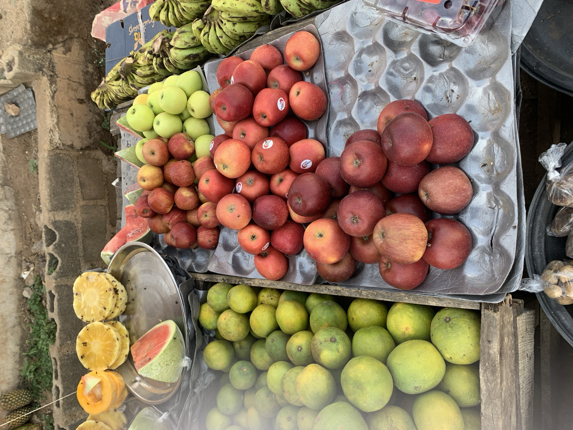 A fruit stand in New Stadium, Nigeria, displays a variety of fruits. Bananas and green apples are at the top. Below them, different varieties of red apples fill two metal trays. A silver platter to the left holds pineapple slices and a watermelon wedge. In the lower part of the frame, green oranges fill a wooden crate. The ground is partially visible to the left, appearing rough and uneven.