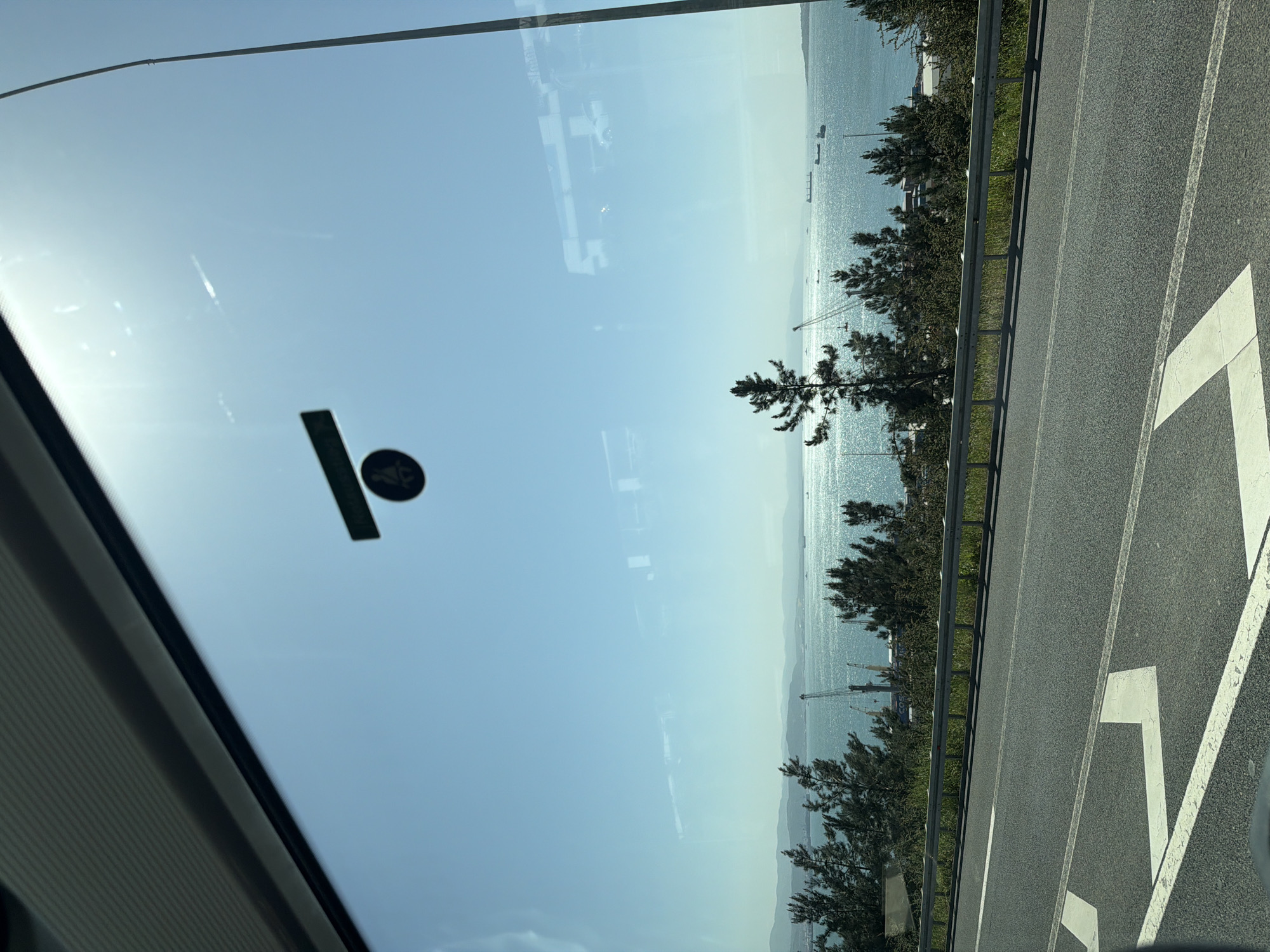 The image captures a sunny daytime view from inside a vehicle, looking out at a coastal highway and a sparkling body of water. The scene is set in Dilovası, Turkey.
In the immediate foreground on the left, the edge of a vehicle's windshield and interior frame are visible. A dark sticker with a circular design and a rectangular bar is affixed to the windshield, catching sunlight. The windshield also shows reflections of what appear to be interior structures or external light sources.
To the right, a multi-lane asphalt highway stretches diagonally, marked with clear white lines, including a large, partially visible numeral "7" or an arrow. A metal guardrail separates the highway from a narrow strip of green grass and a line of dark green, coniferous-like trees.
Beyond the trees, a vast body of water, likely the Gulf of Izmit, glistens intensely under the bright sun, creating a shimmering effect. Several large ships or cargo vessels are visible in the distance across the water. The horizon features hazy, distant land or mountains, indicating a wide expanse. Some industrial cranes or structures are faintly discernible along the distant shore. The sky above is a clear, light blue, with a strong sun flare evident in the upper left quadrant of the windshield.
The overall impression is of a scenic drive along a coastal road on a clear, bright day. No distinct text is readable in the image.