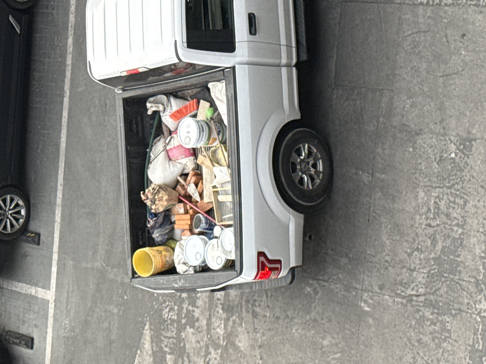 A silver pickup truck, its bed filled with assorted items including buckets, paint cans, bags, and bricks, is parked in a parking lot in San Bernardino Tlaxcalancingo, Mexico. A dark sedan is partially visible to the left of the truck. The parking lot surface is concrete.