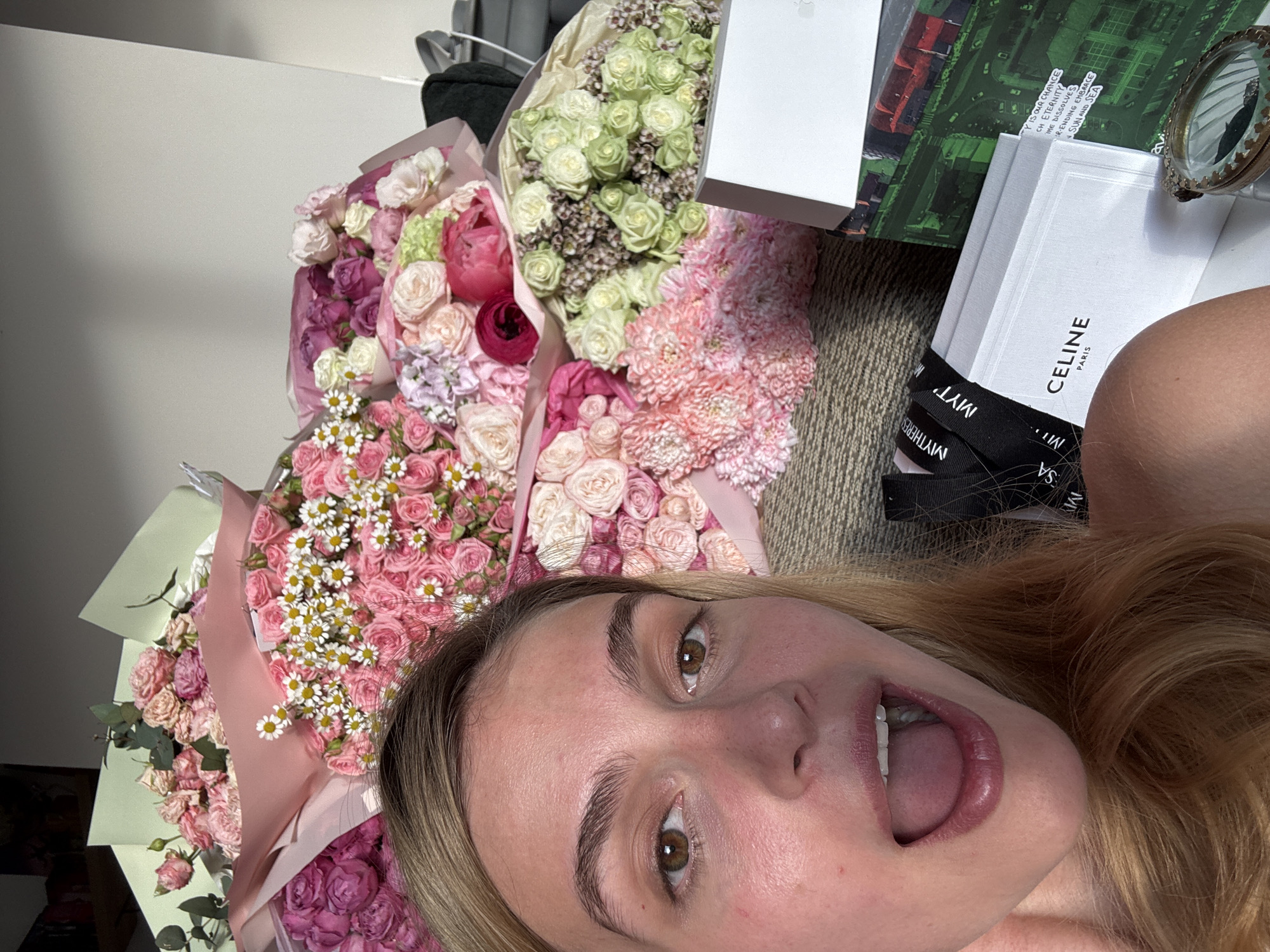 A selfie taken from a low angle, featuring a woman with light skin, light brown hair, and visible brown eyes, mouth agape. She is surrounded by several bouquets of flowers in pink, white, and green hues. Adjacent to the flowers are white boxes, one labeled "CELINE PARIS," and another featuring text including the phrase "Y IS OUR CHANCE." The background shows a textured surface, likely a couch or similar seating area.
