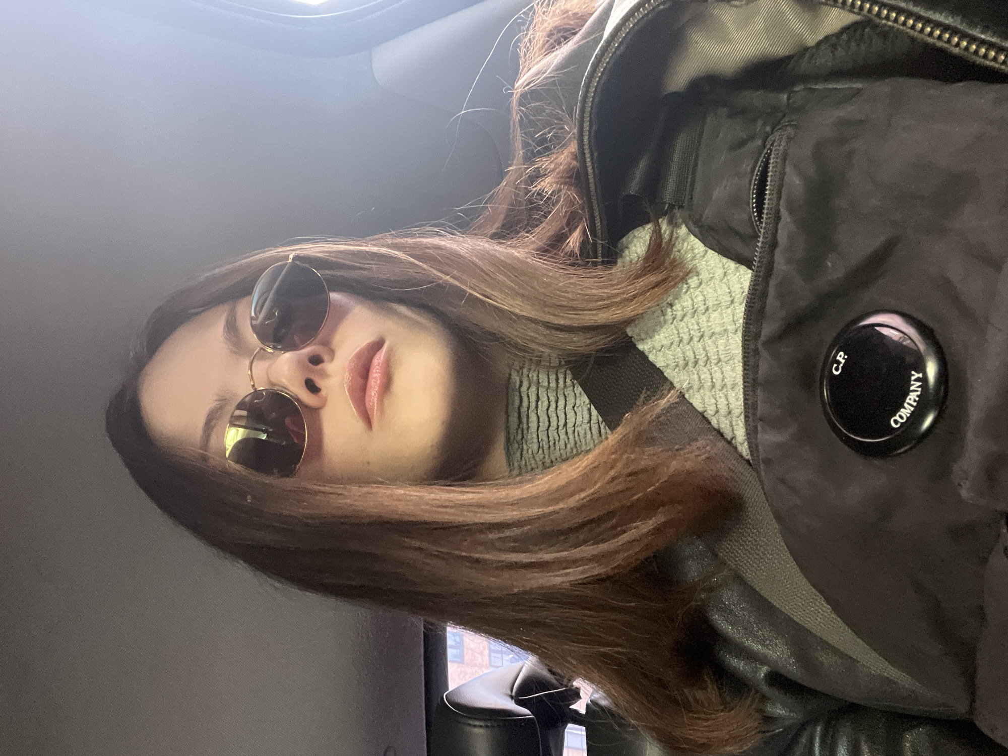 A selfie taken from a low angle features a woman with long, brown hair wearing sunglasses. She is wearing a gray, knitted turtleneck and a black jacket with a "C.P. Company" logo on the sleeve. The environment suggests she is inside a vehicle, possibly a car, with a glimpse of buildings visible through the window behind her. The location is likely in a city, but London cannot be confirmed without further contextual clues.