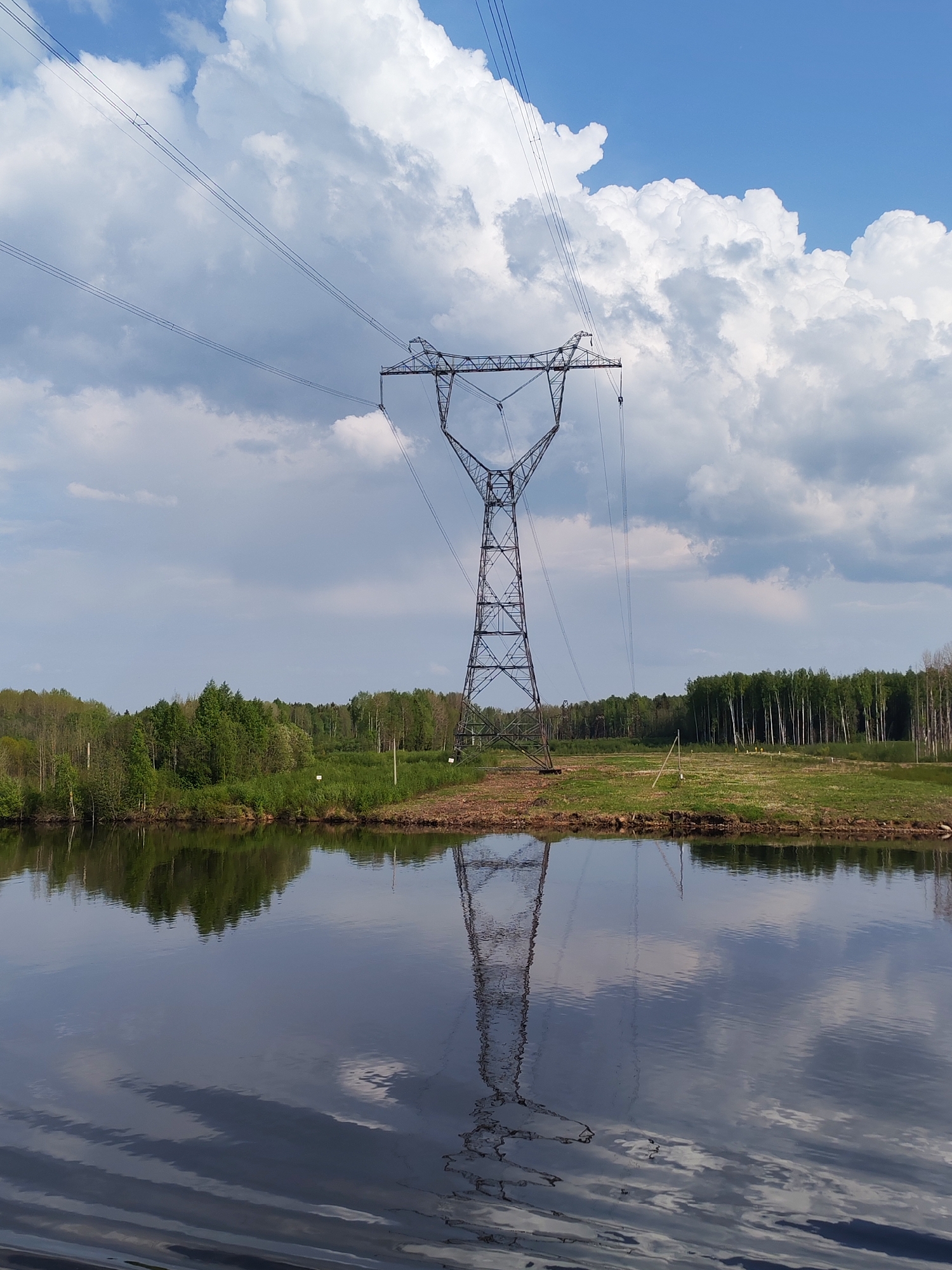 The image depicts a tall, metal electrical transmission tower standing on the edge of a body of water, possibly a river or a lake, with a reflection in the water. The landscape includes forested areas on the opposite bank of the water. Overhead power lines are visible extending from the tower. The sky is partly cloudy with varying shades of blue. Based on the prompt specifying "Gari, Russia," this is likely the location of the scene. The photograph focuses on the power transmission structure and its setting within a natural environment.