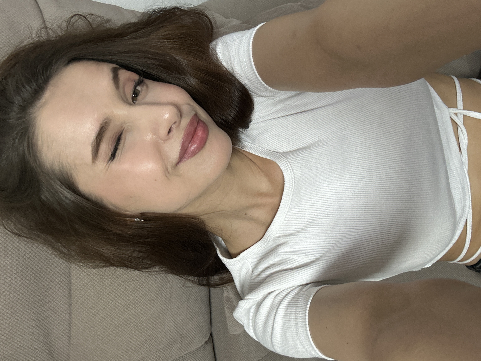 A young woman with brown hair is lying on a beige sofa. She is wearing a white ribbed t-shirt with straps visible on her right side. She is winking with her left eye and smiling. The image appears to be taken at a close range. There is no indication that the image was taken in the city of Yekaterinburg, Russia.