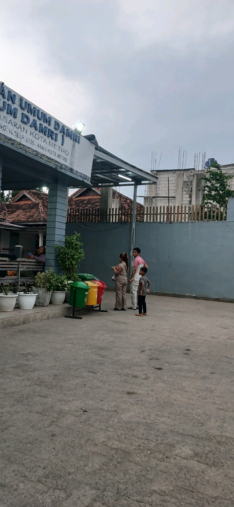 The image depicts the entrance area of an office building, likely a public transportation terminal, indicated by the signage that reads "AN UMUM DAMRI". Below this, further text specifies "PASARAN KOTA METRO" and contact information, confirming the location as Metro City, Indonesia.
In the foreground, a grey concrete surface serves as the ground. To the left, a series of white and grey potted plants are arranged along a raised walkway. A person is seated on a wooden bench on this walkway, looking at a phone. Adjacent to the walkway, three colorful trash bins – green, yellow, and red – stand on a black metal frame.
To the right of the trash bins, a group of four people are visible. A woman wearing a light brown jumpsuit stands in profile, holding a phone. Beside her, a man in a pink and white polo shirt and light-colored pants looks forward. A young boy in a grey t-shirt and black pants stands slightly apart, facing the viewer. Behind them, another person, partially obscured, is also present.
The building facade features a grey overhang supported by columns. The sky above is overcast, suggesting a cloudy or possibly rainy day. In the background, traditional tiled roofs of houses are visible, along with a wooden fence and a partially constructed building with exposed bricks and rebar.