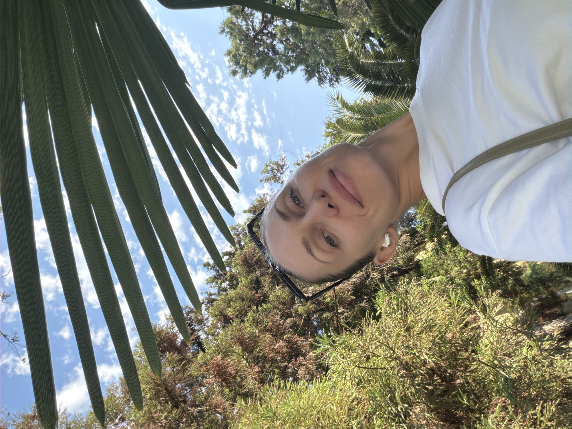 The media file is a portrait-style selfie taken outdoors, possibly in Sochi, Russia, given the foliage and bright sky. The subject is a woman with blonde hair, wearing glasses and a white t-shirt. She has an earbud in her left ear. The photo is taken from a low angle, looking upwards towards the sky and the woman's face. Large palm leaves are in the foreground, creating a frame for the shot. Behind her, there are trees and a bright blue sky with scattered white clouds, suggesting it is daytime with good weather. The woman's expression is relaxed and friendly, with a slight smile. The overall mood is casual and summery. There is no visible text in the image.