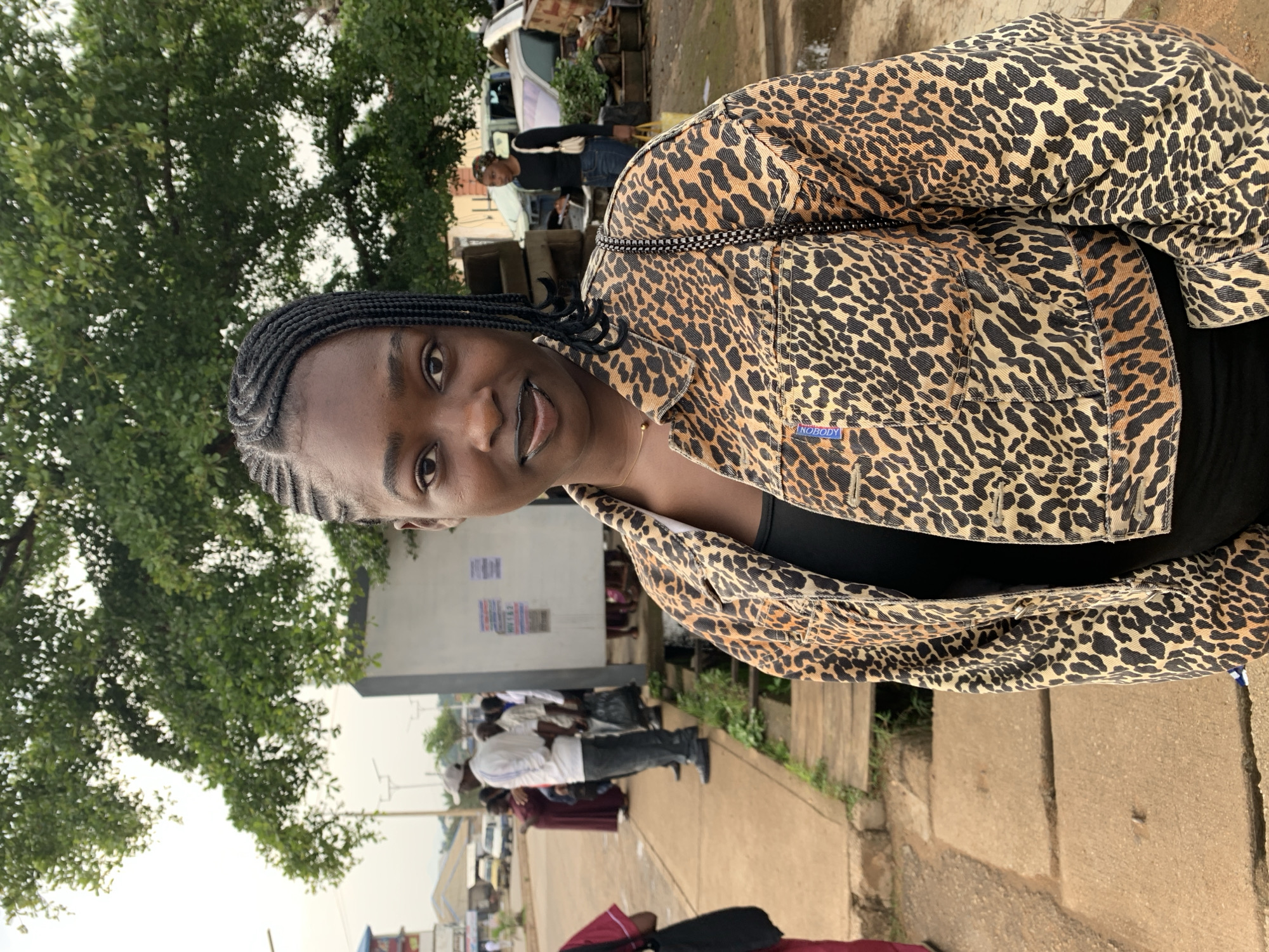 A young Black woman is captured in a medium shot, standing outdoors and facing the camera. She has dark skin, braided hair styled with a center part and braids pulled back, and is wearing dark lipstick. She is dressed in a black top and a leopard-print denim jacket with a subtle blue tag on the left chest pocket. Her body is angled slightly to the left, and she holds her head straight, looking directly at the viewer.
The background suggests an urban environment. To her left, a concrete sidewalk and a drainage channel with wooden planks are visible. Further back, several people are standing and walking, some appearing to be waiting or conversing. A light-colored structure, possibly a building or a bus stop shelter, stands behind them. A large tree with green foliage overhangs the scene from the top left. To the right of the woman, the background shows more of the urban landscape, including what appears to be a parked vehicle and some scattered objects, possibly indicating an informal marketplace or storage area. The overall lighting suggests it is daytime, with diffused light, possibly indicating an overcast sky or the late afternoon.