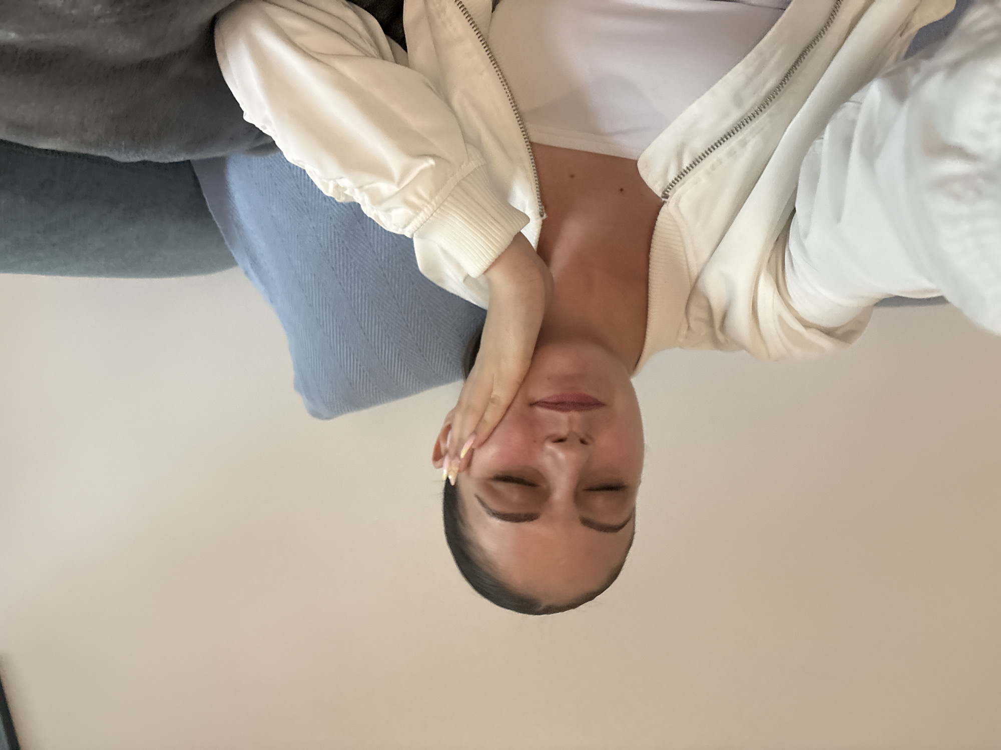 A person is reclining on a couch with their eyes closed. They are wearing a white shirt and a white jacket. Their left hand is resting on their cheek. The couch has a grey cushion and a light blue cushion. The background is a light beige wall. The location in the city of Burgas, Bulgaria cannot be determined from the image.