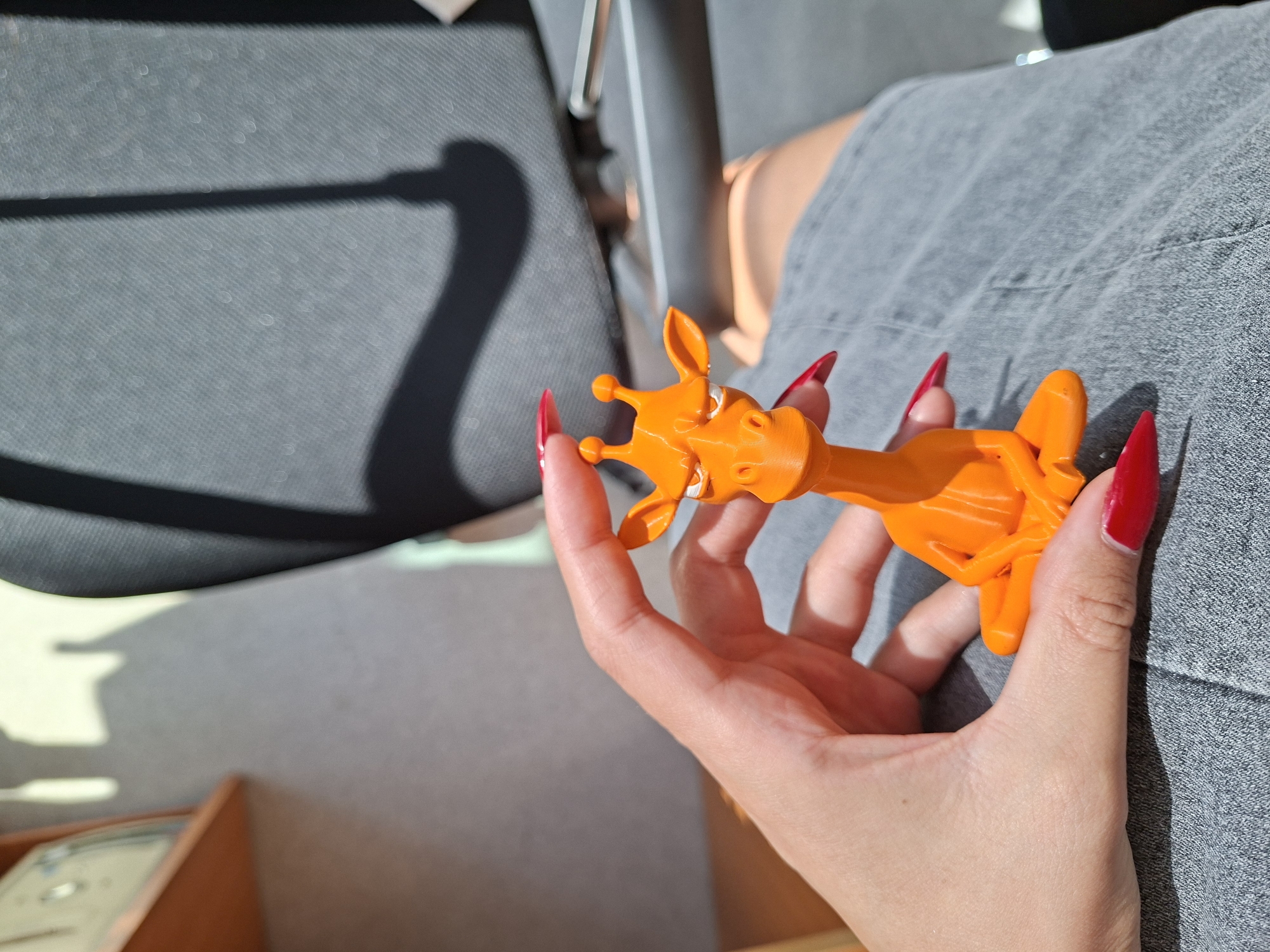 A person's hand with bright red, long, pointed fingernails holds an orange, 3D-printed figurine of a giraffe in a seated, meditative pose. The figurine is small enough to fit comfortably in the hand. The person is wearing grey denim shorts. The background shows the dappled shadow of a chair cast by sunlight, and a blurred wooden shelf. The image appears to be taken indoors, possibly in an office or home environment, with sunlight streaming in from a window.