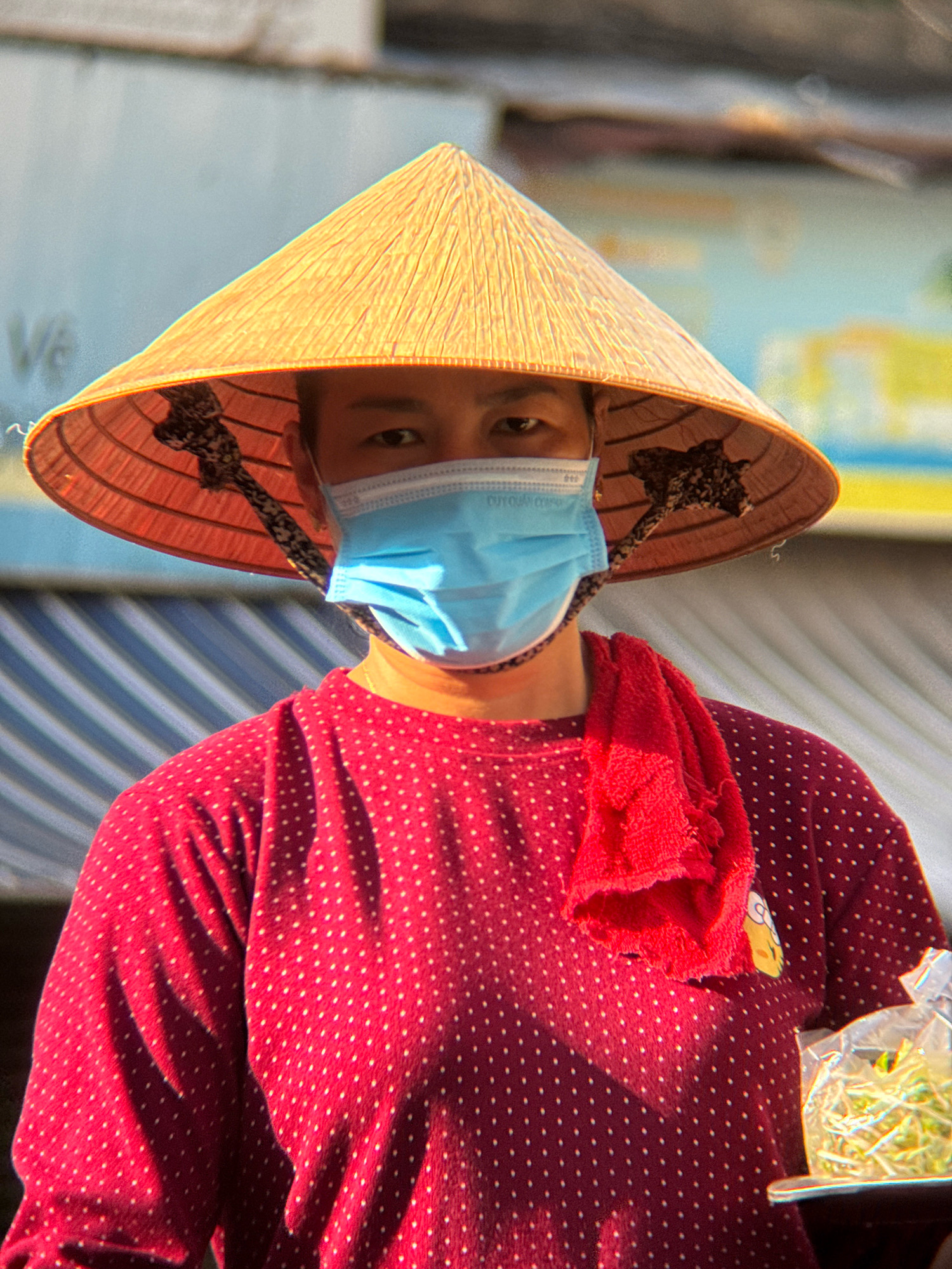 A person wearing a conical hat and a blue face mask stands in a market, likely in Nha Trang, Vietnam. The person is wearing a red polka dot shirt with a red towel draped over their shoulder. In their right hand, they are holding a clear plastic bag containing green sprouts, possibly for sale. The background suggests an outdoor market environment with blurred structures and a striped awning. The lighting indicates it is daytime, with shadows cast on the person's clothing.