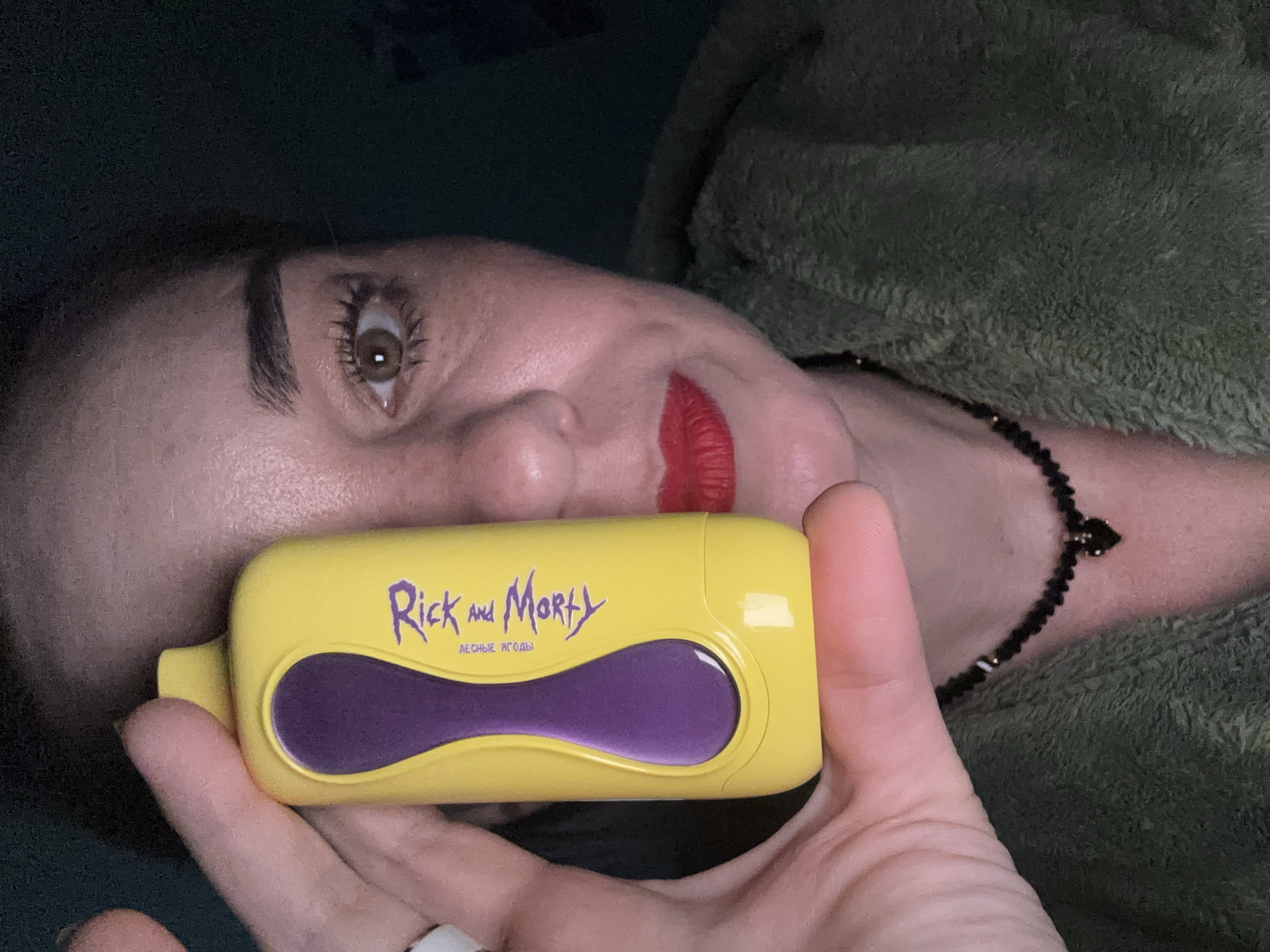 A person is holding a yellow and purple Rick and Morty vaping device in their hand, close to their face. The person has fair skin, brown eyes, and red lipstick. They are wearing a green textured garment. A black beaded necklace is visible on their neck. The background is dark, suggesting an indoor setting. No definitive information to suggest location in the city of Vsevolozhsk, Russia is visible.