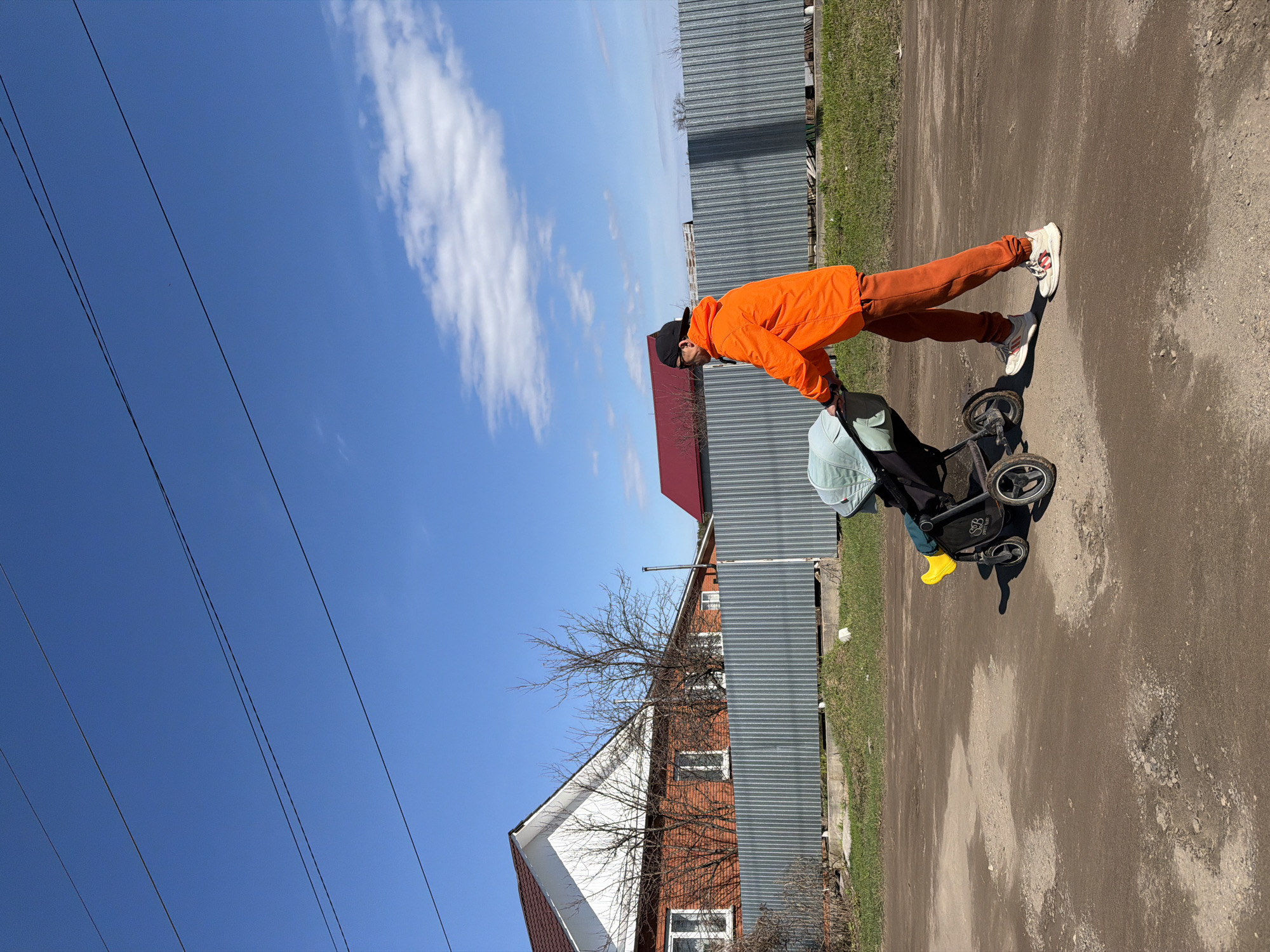 A man in an orange jacket and trousers is walking on a dirt road, pushing a stroller. He is wearing white sneakers and a black cap. The stroller contains a baby seat with a light green cover. A pair of yellow boots are visible in the stroller.
Behind the man, there is a house with a red roof and a gray corrugated metal fence. Power lines are visible in the blue sky. The location is likely Chaikovsky, Russia, given the provided context.