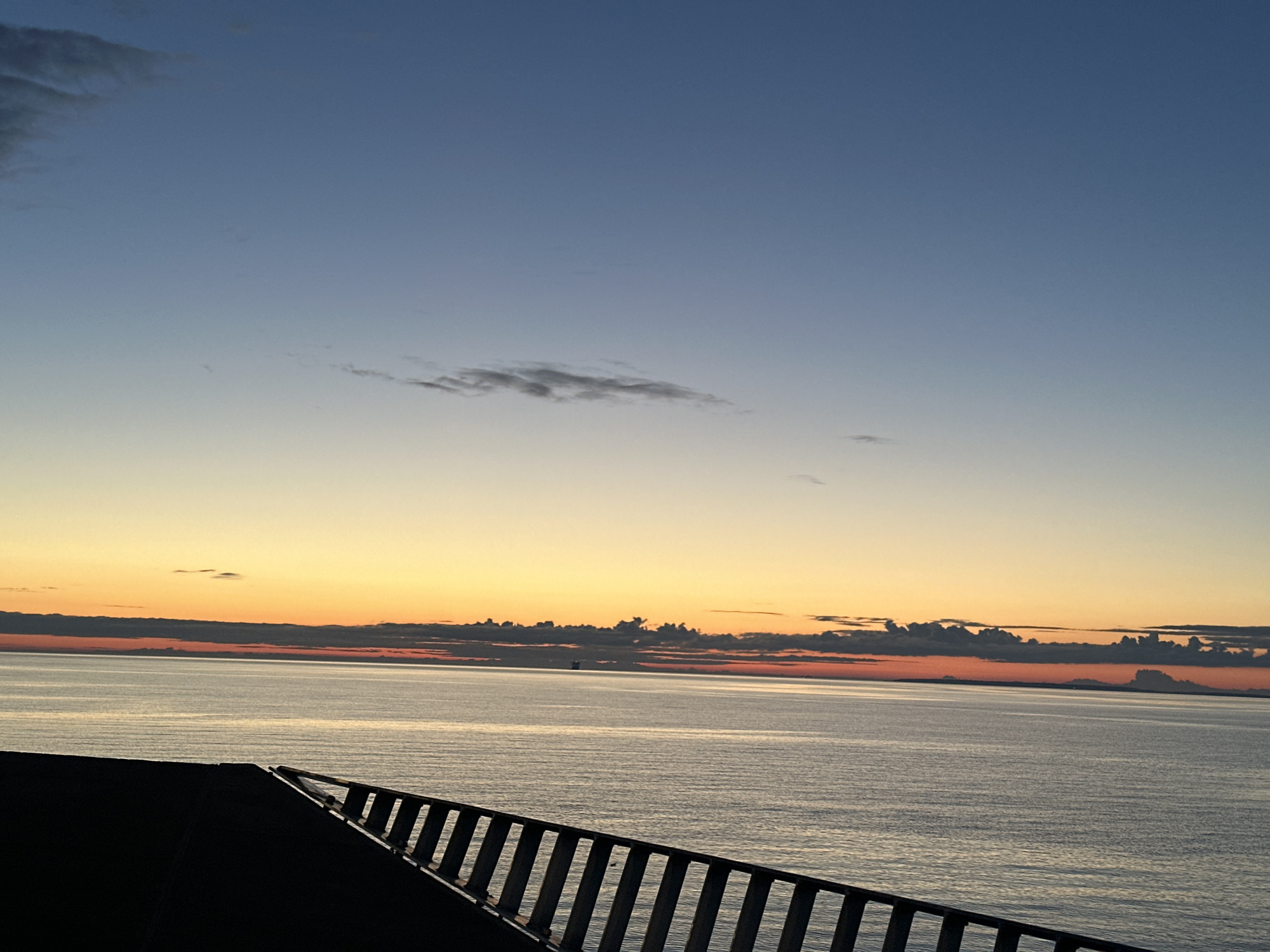 The image shows a serene seascape at dusk, likely taken from an elevated vantage point such as a balcony or a deck overlooking the Baltic Sea. The sky transitions from a deep, clear blue at the top to soft oranges and pinks near the horizon, indicating either sunrise or sunset. Several wispy clouds are scattered across the sky.
The sea is calm, reflecting the muted colors of the sky. In the distance, a faint silhouette of a structure, possibly a buoy or a distant vessel, can be seen on the water. Along the bottom of the image, a dark, angled structure with a railing is visible, suggesting a man-made observation area or the edge of a ship. The railing consists of vertical metal bars.
While the image captures a beautiful natural scene, it does not contain any specific elements that allow for definitive identification of the location within Tallinn, Estonia. However, given the seascape and the likelihood of being near the coast, potential viewing locations could include areas along the Tallinn Bay, such as Pirita or Kalamaja, or from a ferry or cruise ship docked in the port. The overall atmosphere is peaceful and tranquil.