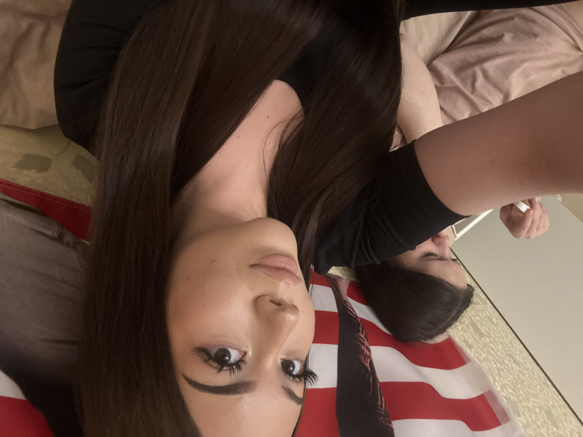 An indoor, vertically oriented image features two young women lying down, seen from an overhead perspective. The primary subject, positioned in the foreground and lower left, has long, dark brown hair that cascades around her. She wears a black top and looks directly at the camera with a neutral expression, enhanced by long eyelashes and light makeup. Her face is well-lit, highlighting her features.
A second woman is partially visible in the background on the right side of the frame, also with dark hair and wearing a black top. Her face is mostly turned away or obscured, and her right arm is visible.
Beneath the subjects, a large American flag with distinctive red and white stripes is draped, providing a strong visual element in the bottom and middle sections of the image. The rest of the background consists of soft, rumpled bedding or blankets in beige and light pink/peach tones, suggesting a bedroom or relaxed setting. The women appear to be posing for a selfie while relaxing. No specific time of day or weather is discernible, and no text is visible within the image. While the image does not provide direct visual cues for Naberezhnye Chelny, Russia, this is the provided location context.