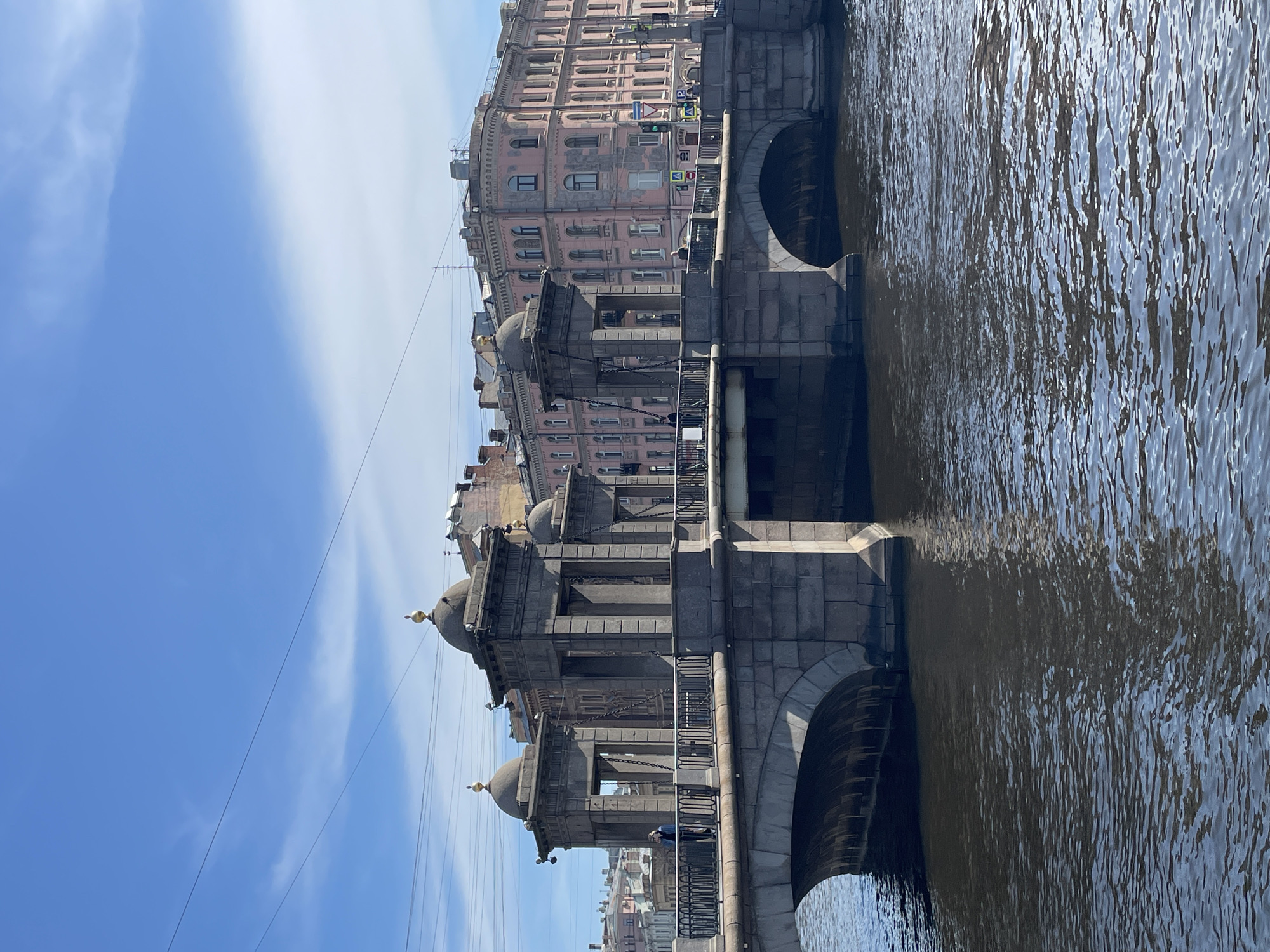 This outdoor scene depicts a section of the Lomonosov Bridge in Saint Petersburg, Russia. The bridge, constructed of grey stone, spans a body of water, with architectural features like arches and decorative cupolas with golden spires visible. Buildings in soft hues, predominantly pink, line the background along the riverbank. The sky is a clear blue with wispy white clouds. The water surface shows ripples, suggesting movement, and reflects the buildings and bridge structure.