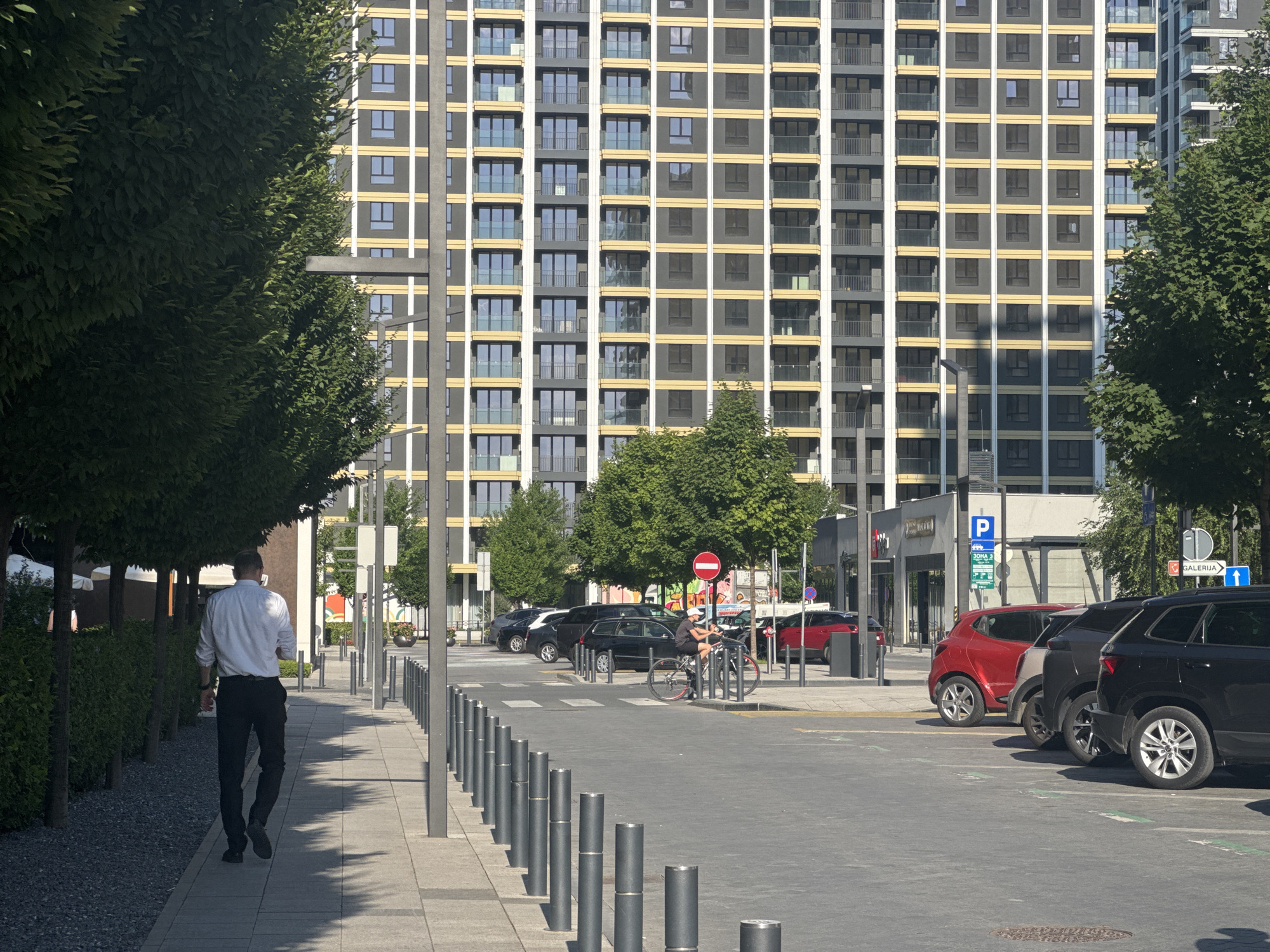 The image depicts a modern street scene in Belgrade, Serbia, characterized by a large residential building and several cars parked along the street. A man in a white shirt and black trousers walks away from the camera on a paved sidewalk lined with trees and planters. In the background, a multi-story building with a grid-like facade of windows and balconies dominates the skyline. Several cars, including black sedans and a red SUV, are parked on the street. A person on a bicycle is also visible near the parked cars. Street signs and bollards are present, indicating a regulated urban environment. The scene is bathed in sunlight, casting shadows on the ground.