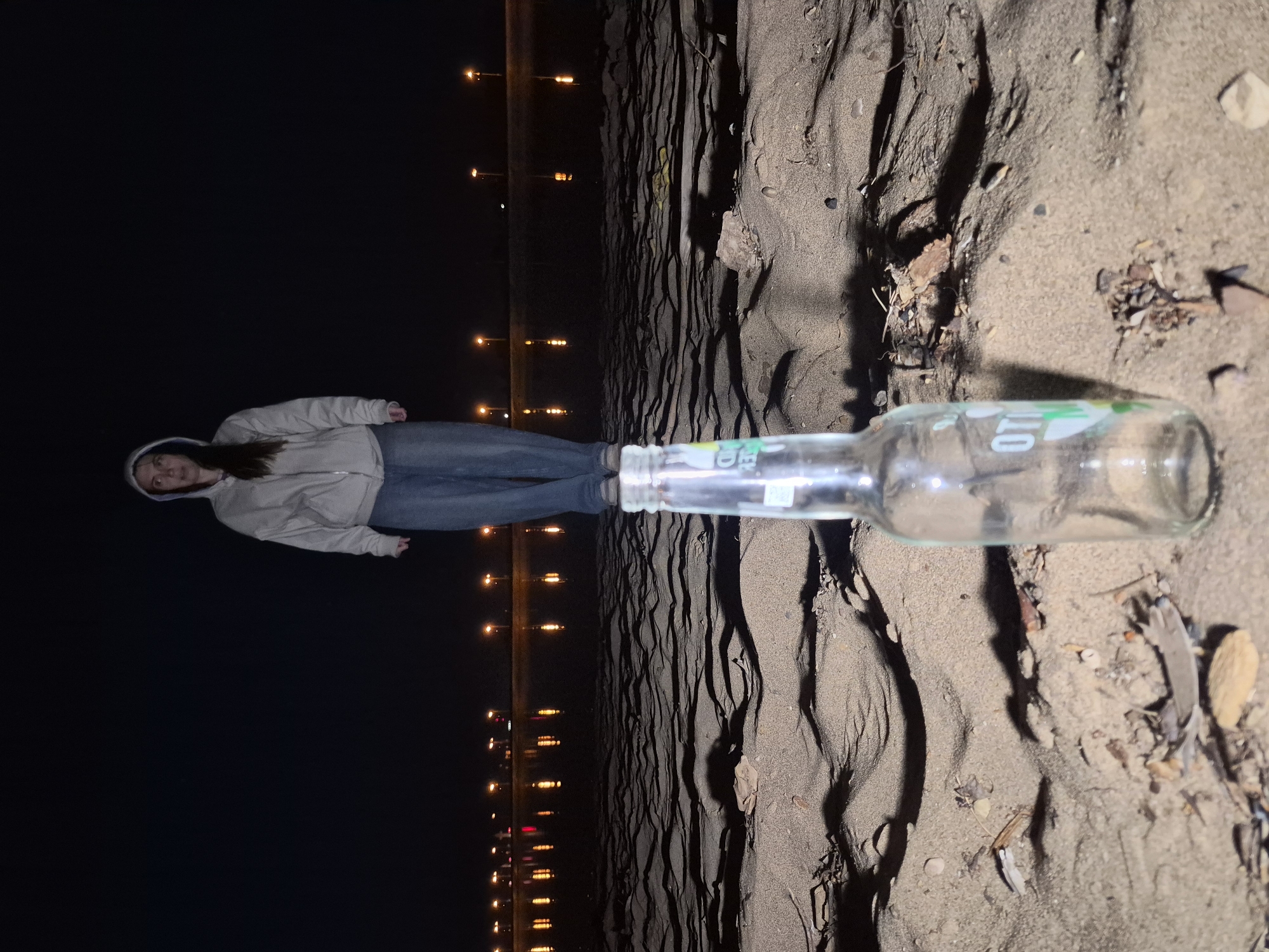 A nighttime scene shows a person, an empty glass bottle, and a sandy area, possibly a beach. The person is positioned on the left, wearing a light-colored hooded jacket and jeans, with a line of distant lights stretching along the horizon behind them. The empty glass bottle lies horizontally across the foreground, seemingly partially buried in the sand. The sand itself is textured with small ridges and scattered debris like leaves. The city is unknown.